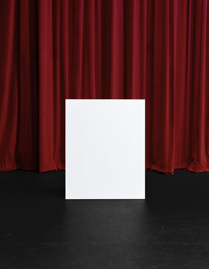 A white rectangular form stands centered on a dark floor against deep red velvet curtains, creating a stark geometric composition with dramatic contrast between the cool white plane and warm burgundy drapery.