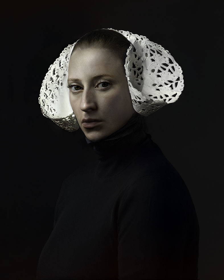 Portrait of a woman against a dark background wearing a black garment and an ornate white lace coif with openwork detailing that frames her face. The dramatic lighting emphasizes the delicate texture of the headpiece while her expression remains composed and contemplative.