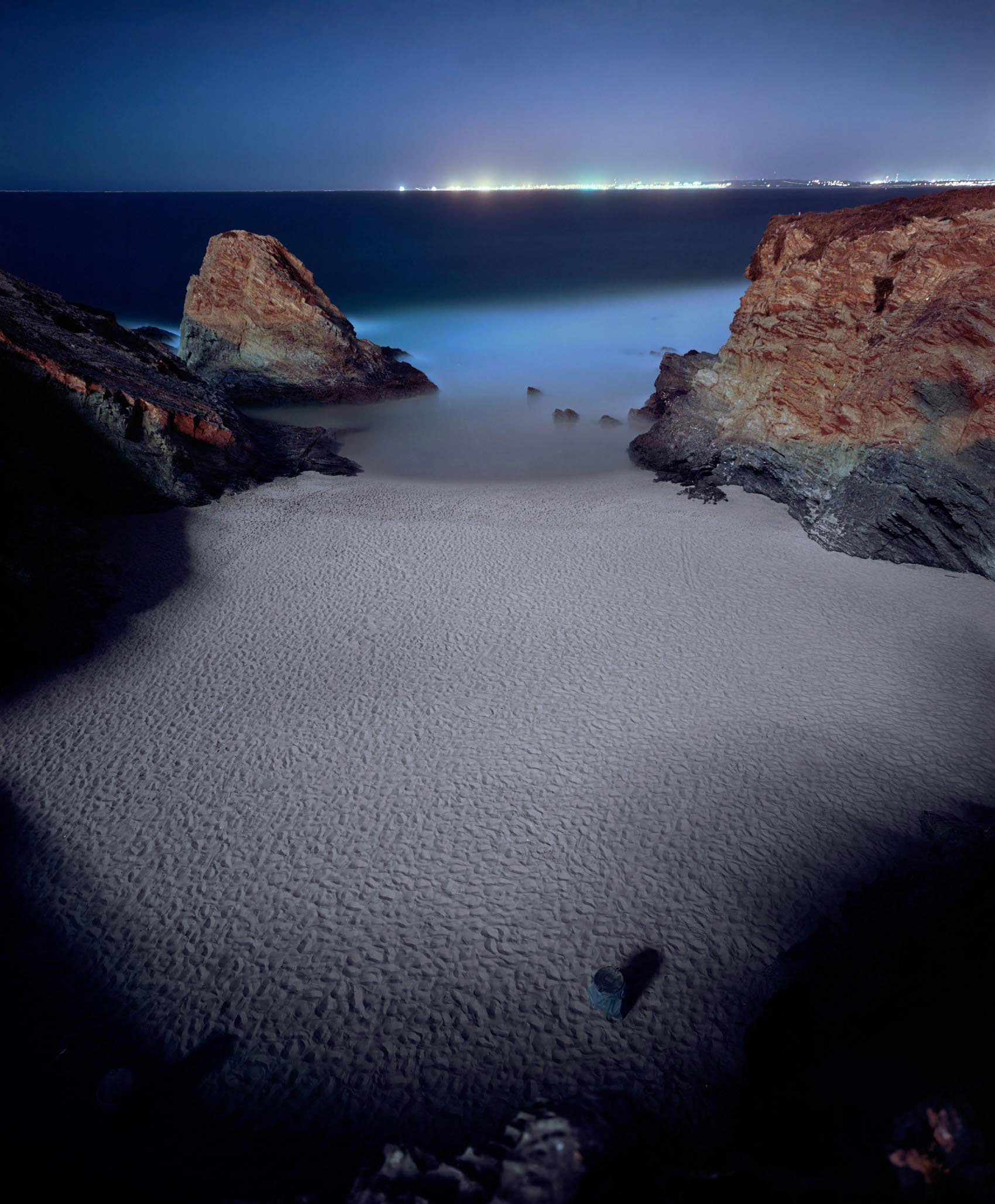 A nocturnal coastal landscape featuring a sandy beach framed by reddish-brown rocky outcrops on either side. The water is rendered in soft, ethereal tones of blue and white, with distant city lights glowing along the horizon. The long exposure technique creates a ethereal, dreamlike quality with smooth water surfaces and subtle texture across the sand.