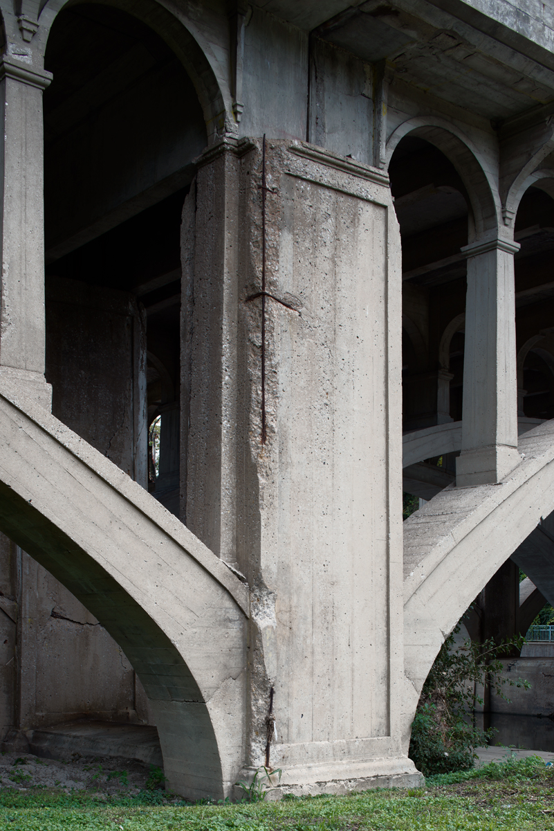 Concrete bridge understructure with massive supporting columns and arched openings, showing weathered surfaces and moss growth. Architectural view emphasizes geometric forms and the monumental scale of the brutalist structure.