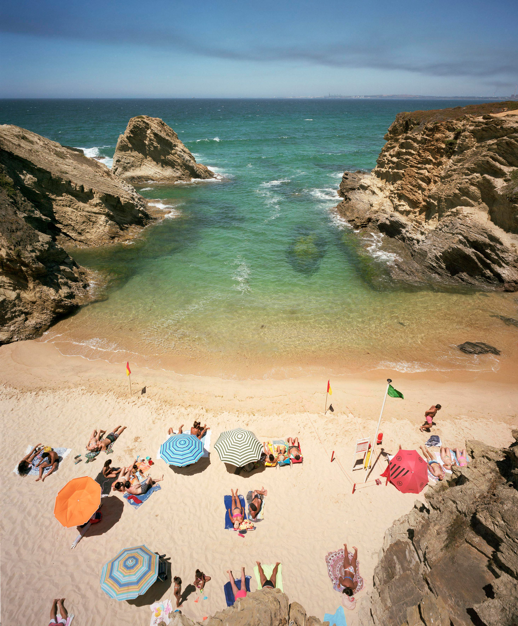 Aerial view of a sandy cove nestled between dramatic rocky outcrops, where numerous beachgoers are scattered across the sand with colorful umbrellas in orange, blue, pink, and striped patterns. Clear turquoise water meets the shore while flags mark sections of the beach, and the horizon extends to a distant shoreline under a partly cloudy sky.