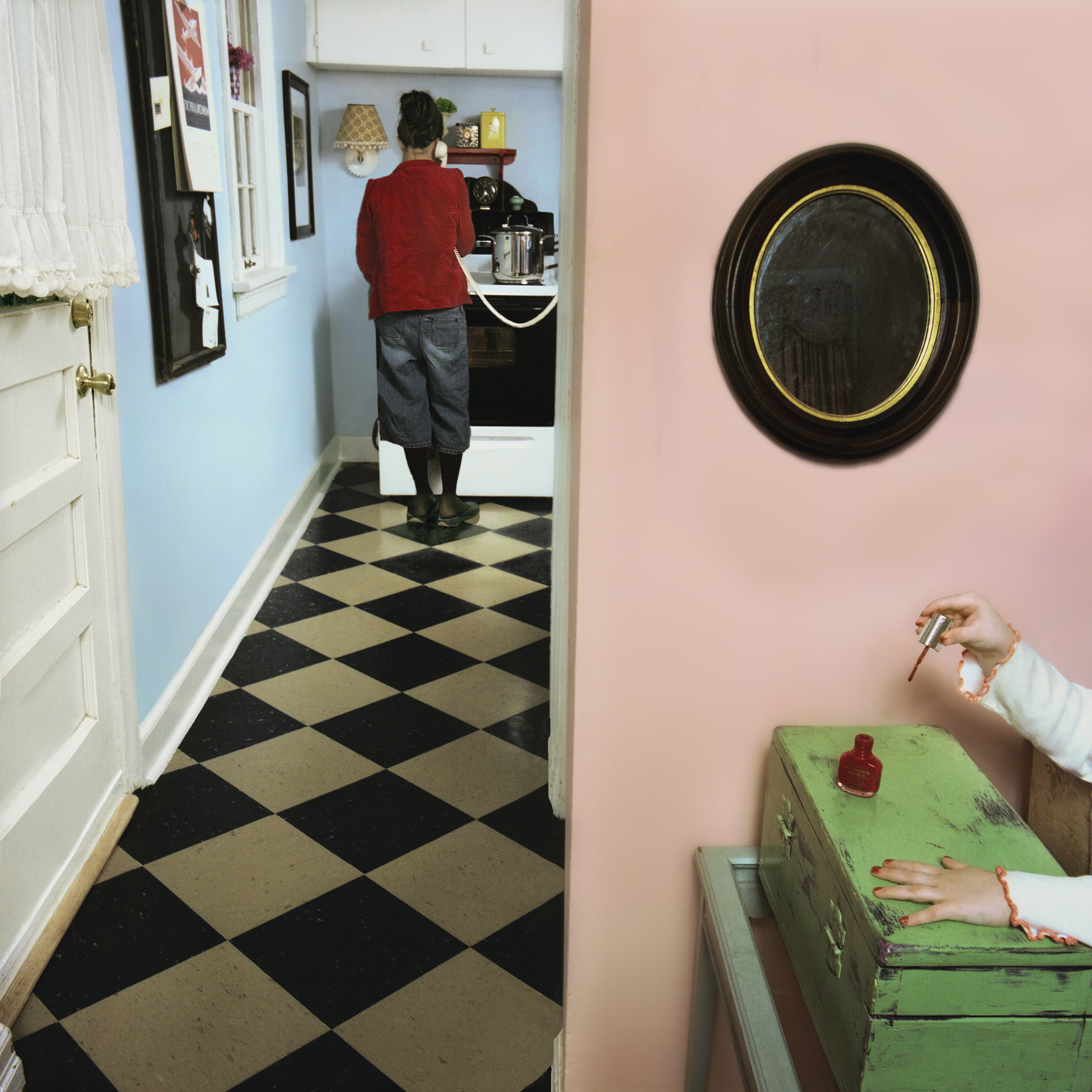 A woman in a red jacket stands in a narrow hallway with black and cream diamond-patterned flooring, facing away toward a kitchenette with blue walls. In the foreground on the right, a person's hand applies nail polish while resting on a distressed green dresser, with an oval mirror and pink wall visible above.
