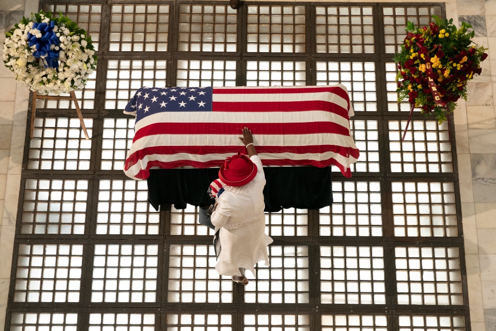Overhead view of a person in white clothing and a red hat standing before a casket draped with the American flag, flanked by two floral wreaths on either side.