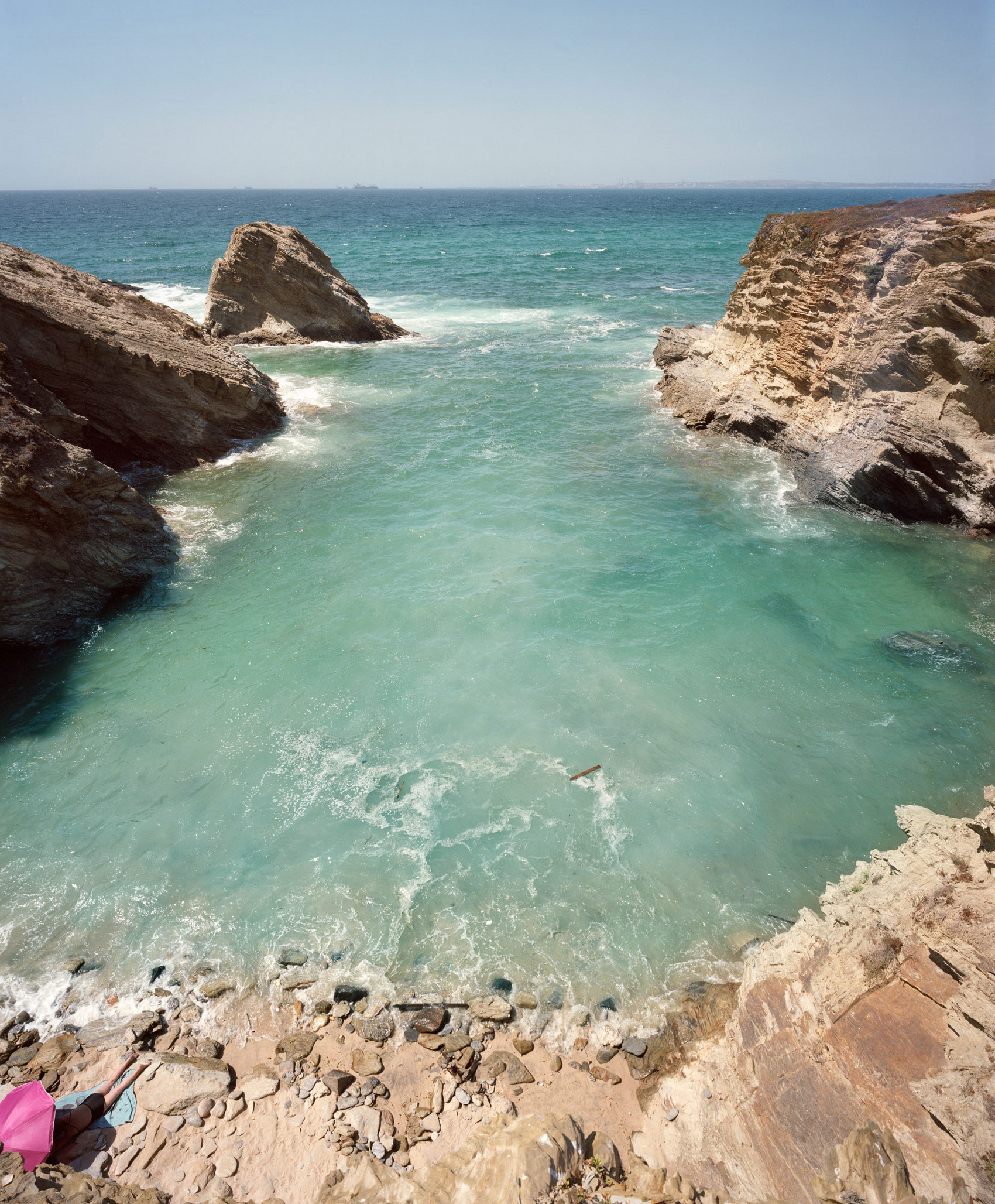 Coastal landscape featuring turquoise waters framed by rocky limestone cliffs, with a solitary rock formation rising from the sea and a small sandy beach in the foreground where a person in pink clothing sits.