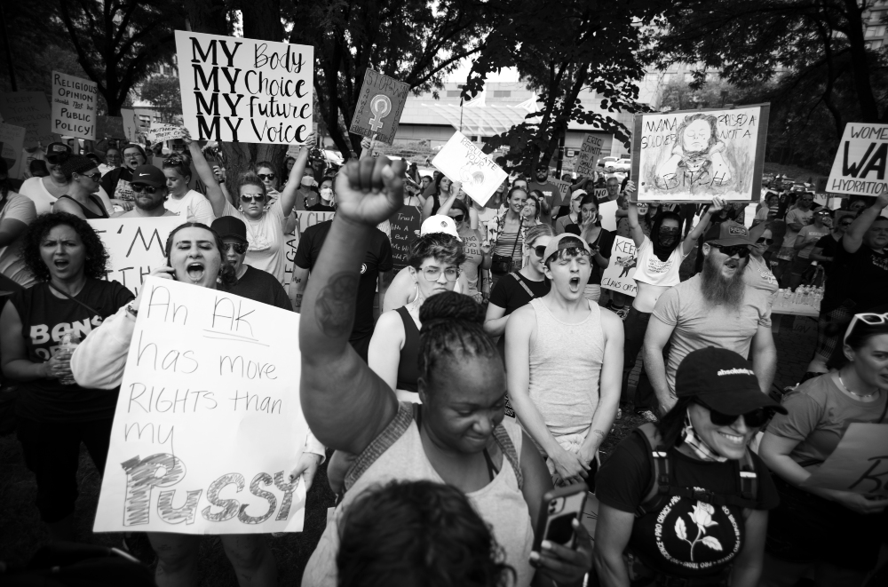 Black and white photograph of a large outdoor protest with dozens of demonstrators holding signs. Visible placards include messages about bodily autonomy and reproductive rights. Participants of various ages and backgrounds are densely gathered beneath trees, with raised fists and expressions of determination and passion visible throughout the crowd.
