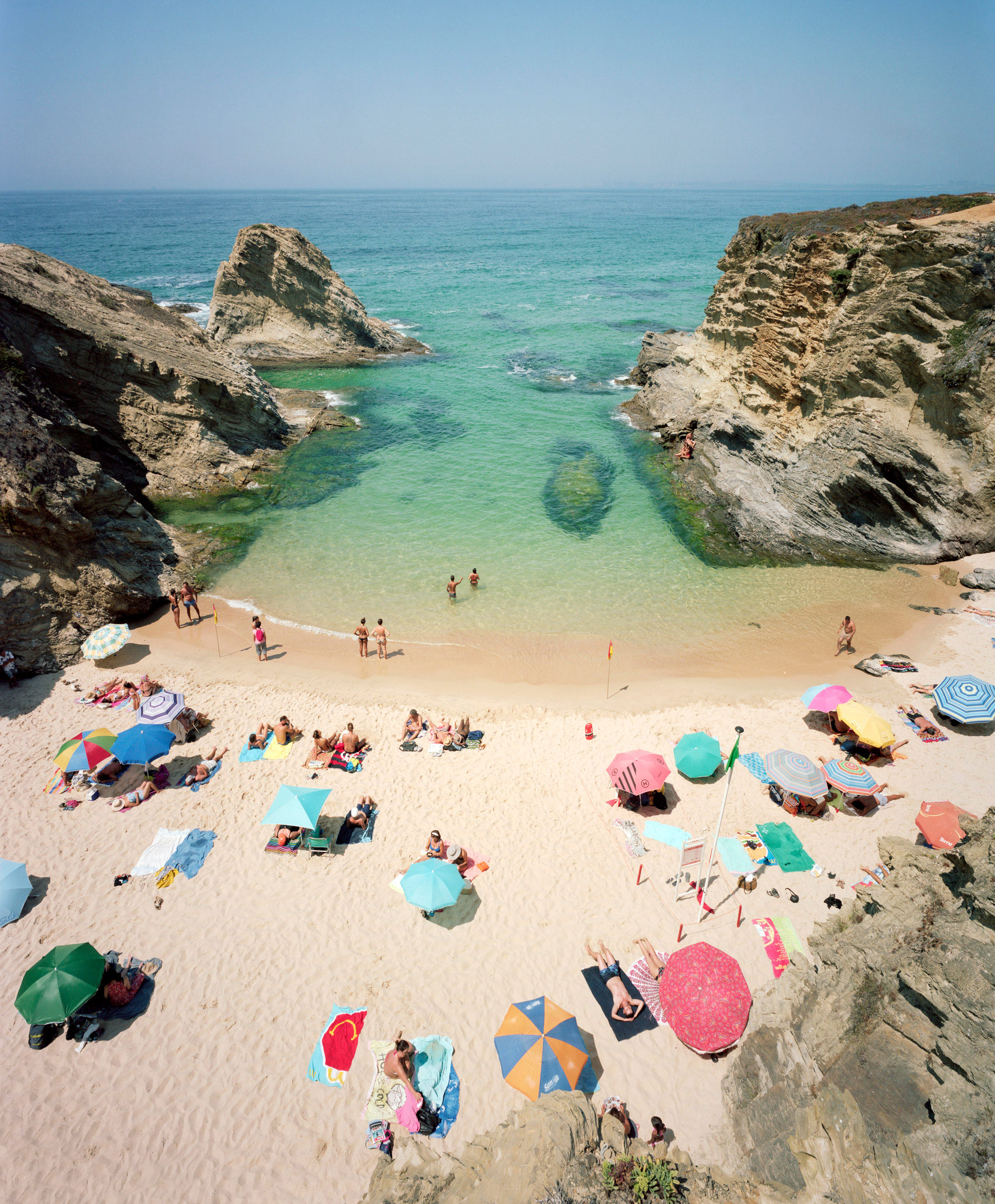 Aerial view of a secluded cove beach framed by dramatic limestone cliffs, with colorful umbrellas and beachgoers scattered across golden sand and turquoise shallow waters.