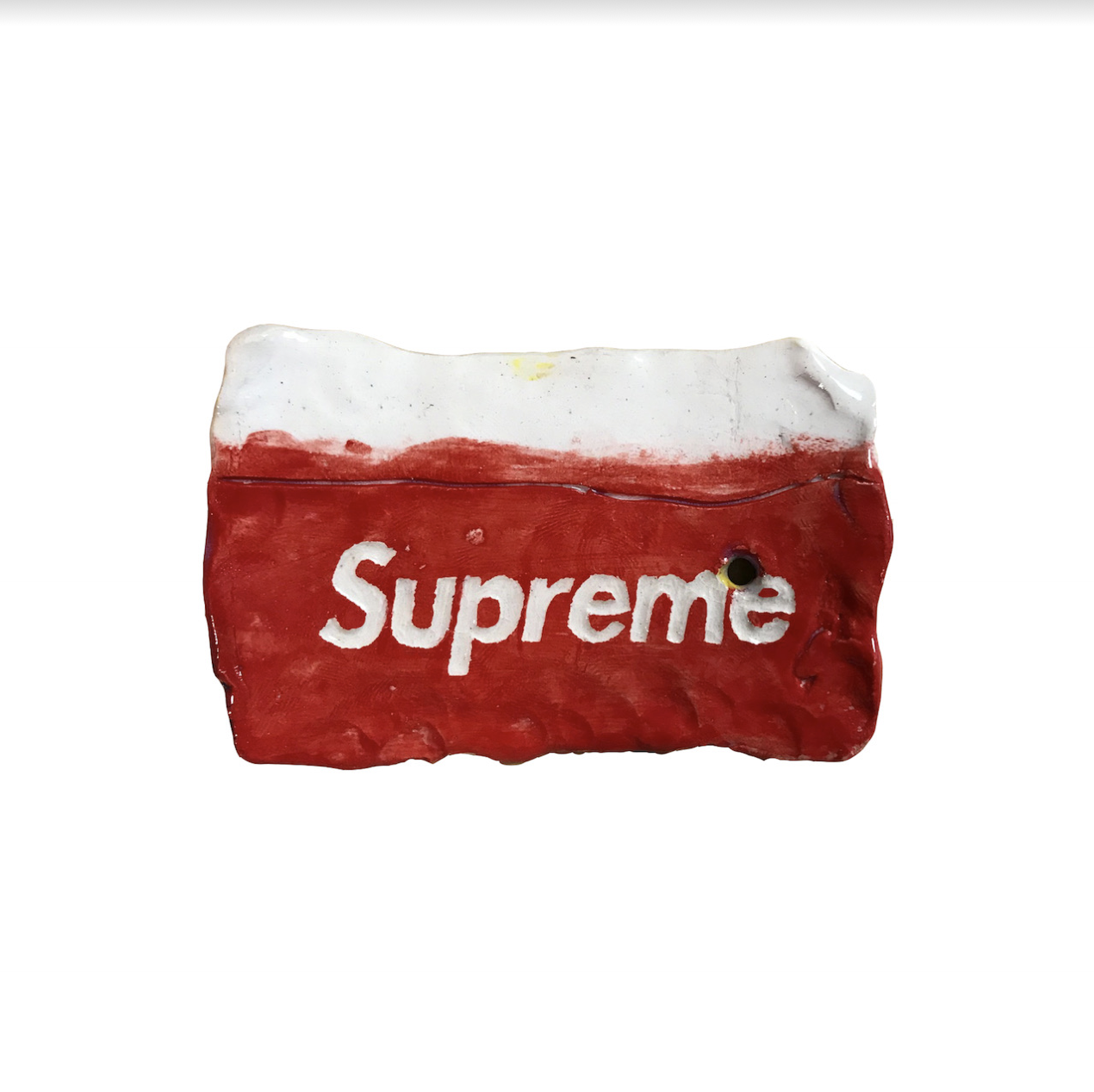 A red and white ceramic object resembling a worn metro card, with the word "Supreme" printed in white across its red surface. The piece is crafted from clay with glazed finish, showing deliberate signs of distress and weathering, with a small hole punctured through one side.