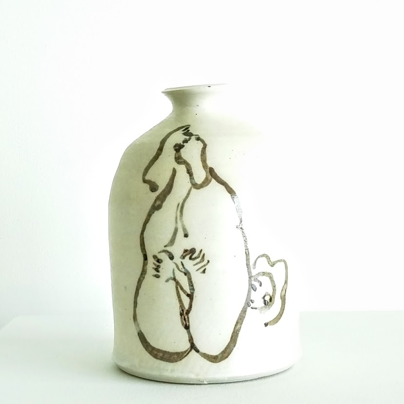 A cream-colored ceramic vessel with a narrow neck and rounded body features a minimalist ink drawing of a reclining nude figure rendered in loose, gestural lines. A small handle extends from the lower right side of the form.