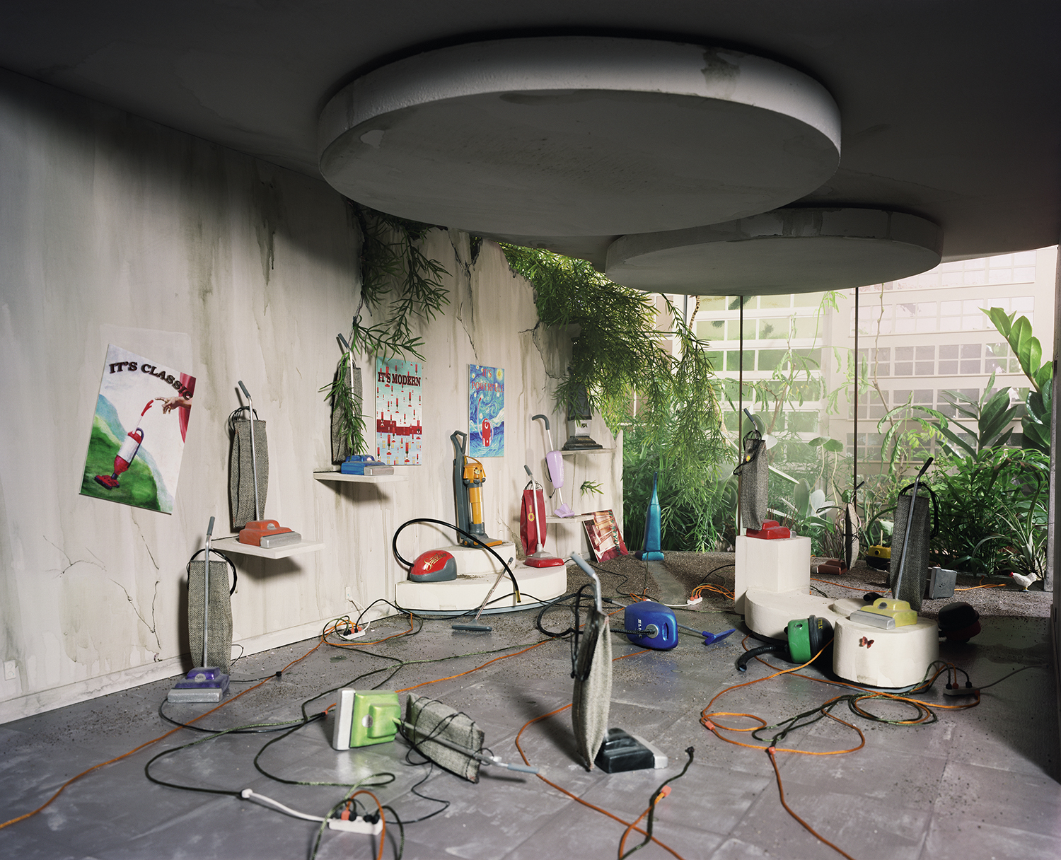 A meticulously constructed interior space featuring vacuum cleaners and cleaning supplies arranged on shelves and pedestals against white walls, with lush green plants hanging from a circular concrete ceiling element and visible through large windows to a garden area beyond. Orange and black electrical cords crisscross the gray floor in an organized display that blurs the distinction between product showroom and immersive installation.