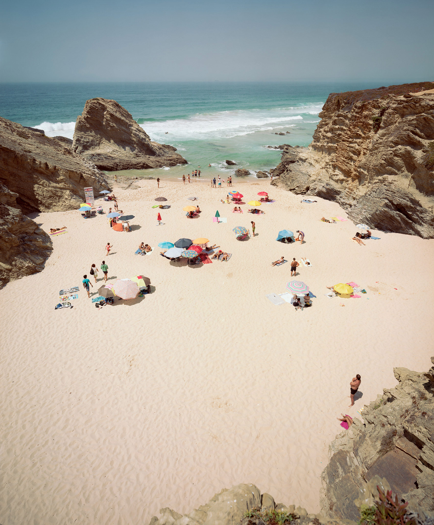 Aerial view of a sandy cove enclosed by dramatic rocky cliffs, with numerous beachgoers scattered across the sand under colorful umbrellas and beach tents, the turquoise ocean and white-capped waves visible beyond.