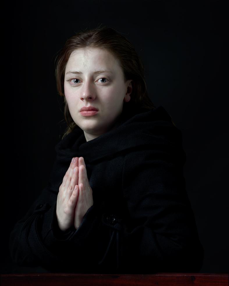 Portrait of a young woman with dark hair against a black background, wearing a black garment with her hand raised to her chest in a contemplative gesture. The stark lighting emphasizes her pale complexion and direct gaze toward the viewer, creating an intimate, introspective mood.