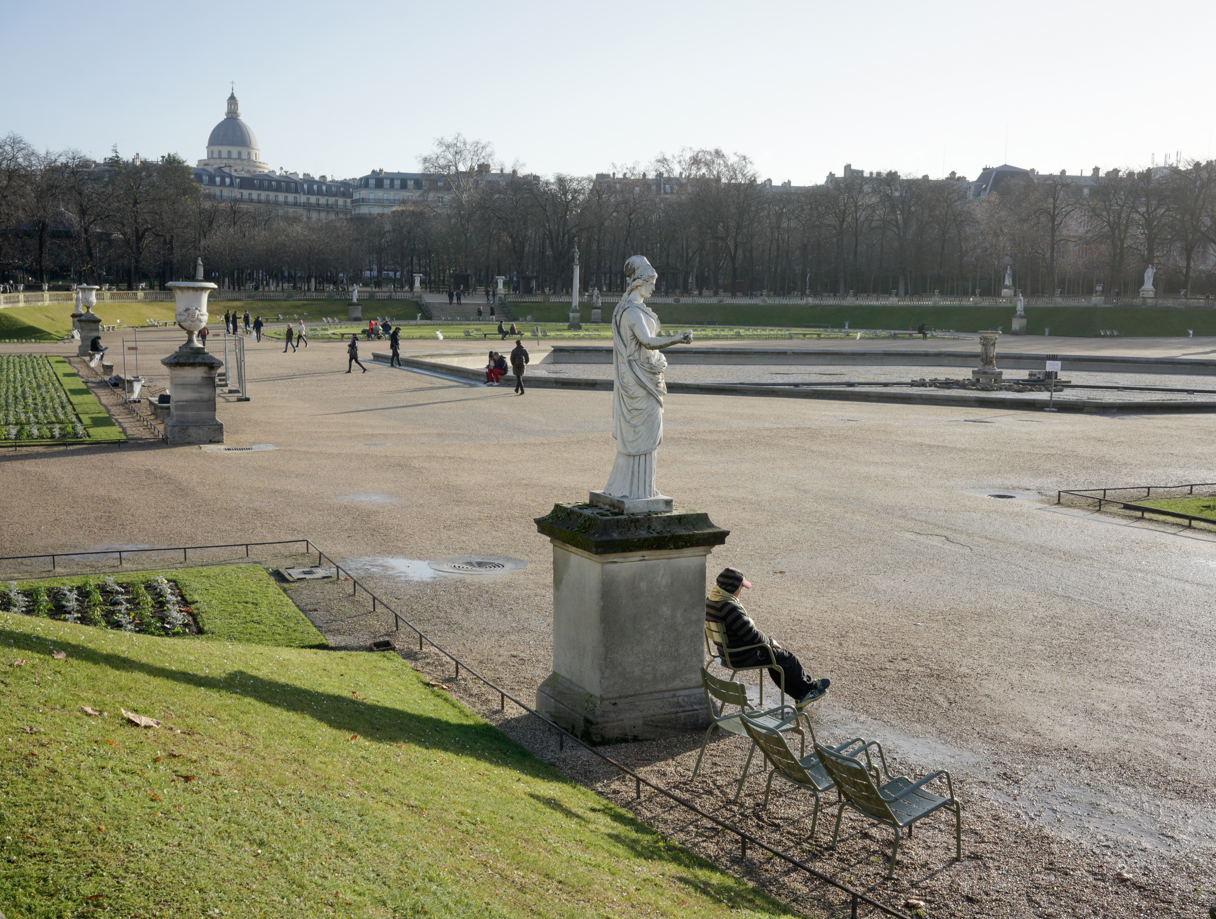 A person sits on a metal chair facing a white classical statue in a vast formal garden. In the background, manicured lawns, rows of bare trees, a geometric water basin, and a domed building create a symmetrical Parisian landscape under overcast light.