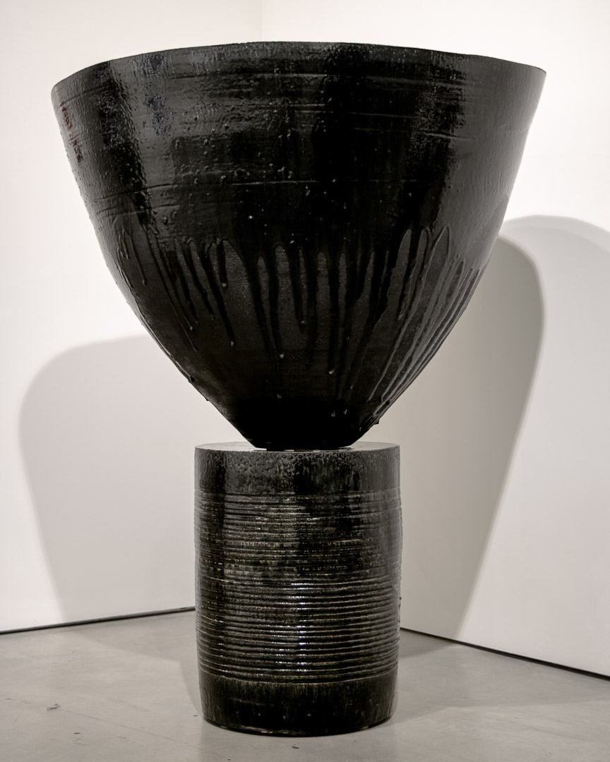 A tall dark wooden vessel composed of two stacked cylindrical forms—a textured base with horizontal incised lines topped by a flared, open bowl with radiating vertical grooves along its lower half. The sculpture casts a dramatic shadow against a white gallery wall.