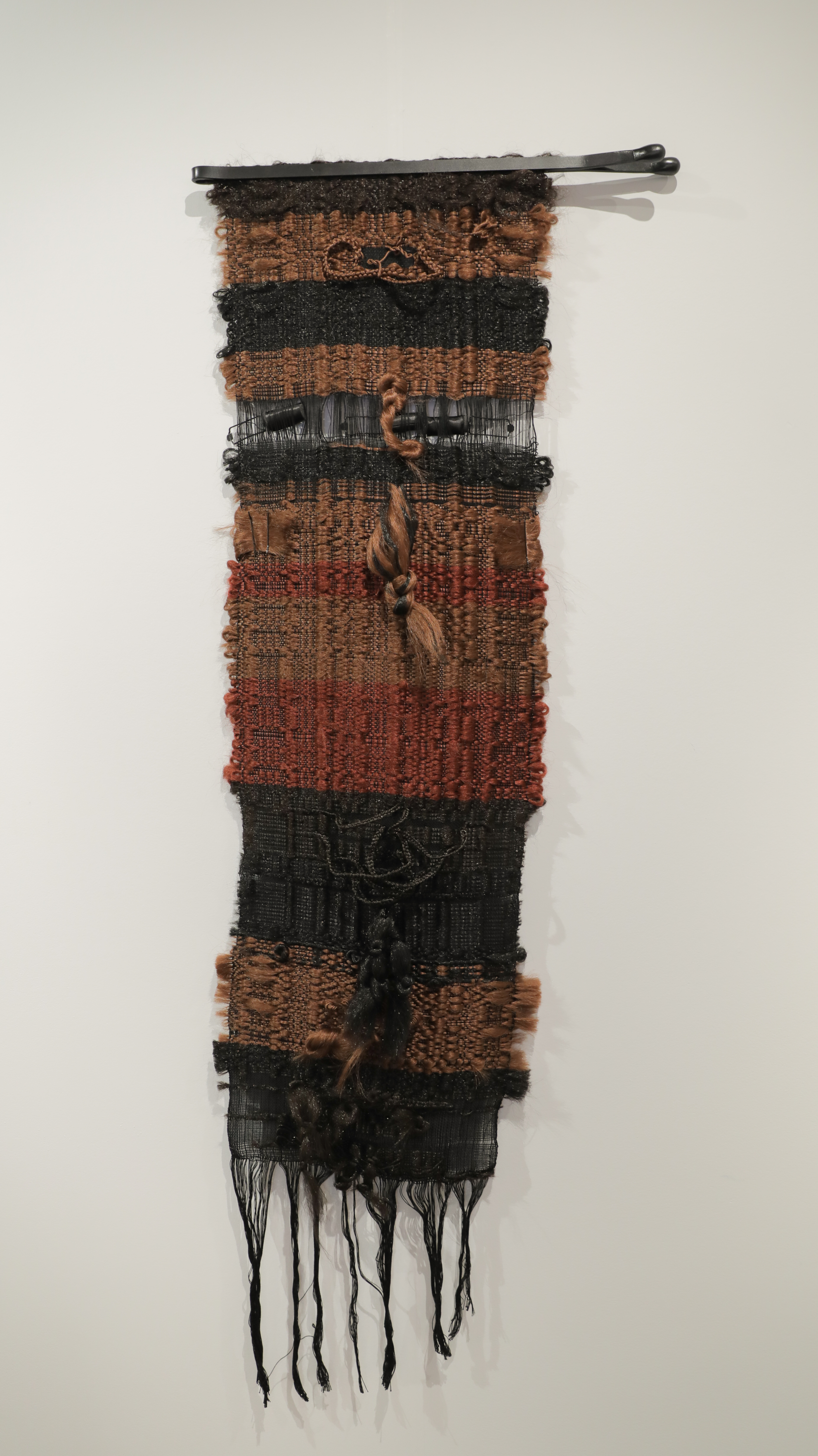 Vertical textile wall hanging composed of alternating bands of woven fabric in copper, rust, and black tones, with synthetic hair woven throughout and secured with satin rollers and bobby pins at intervals. Black fringe extends from the bottom edge.
