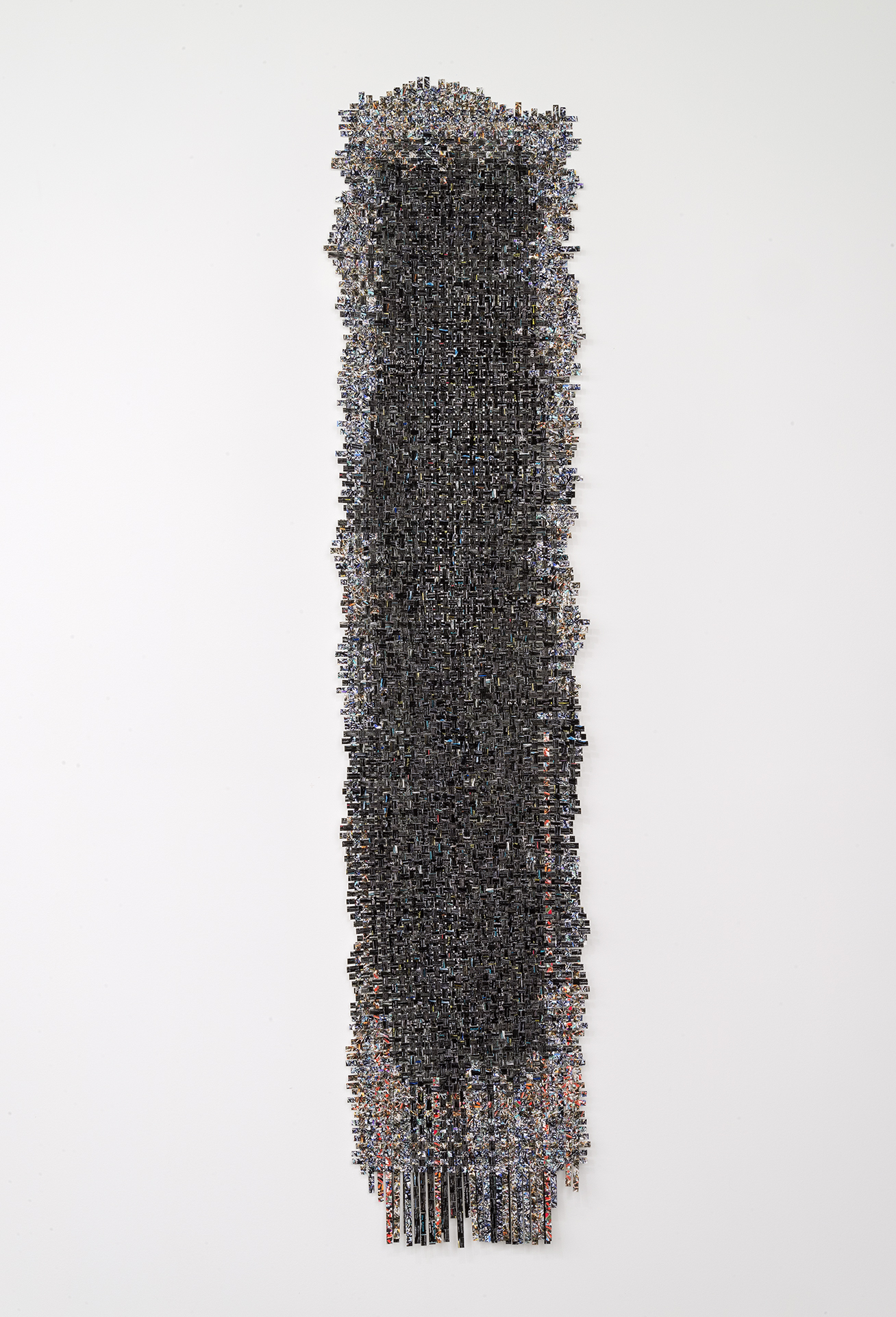 A tall, narrow vertical composition densely composed of hundreds of small rectangular elements in dark grays, blacks, and muted earth tones, arranged in a tightly woven grid pattern. The form tapers slightly at top and bottom, with loose strands or threads extending downward, creating a textile-like appearance against a white background.