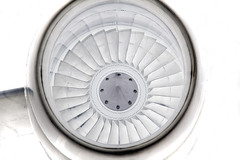 Overhead view of a jet engine's fan blades radiating outward in concentric white metal rings, photographed from directly above. The geometric pattern creates a symmetrical spiral composition with a central circular hub, rendered in grayscale tones.