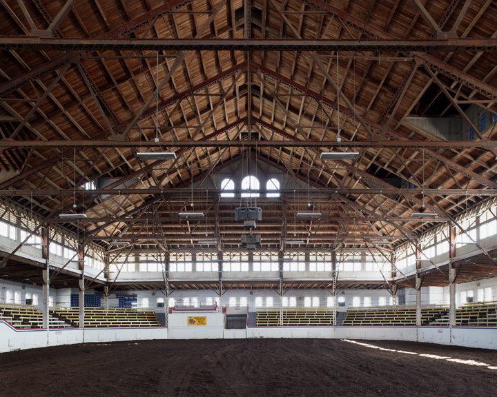 Interior view of a vast wooden-beamed arena with a dirt floor, yellow and white bleachers lining the sides, and natural light streaming through arched windows and skylights in the peaked roof structure.