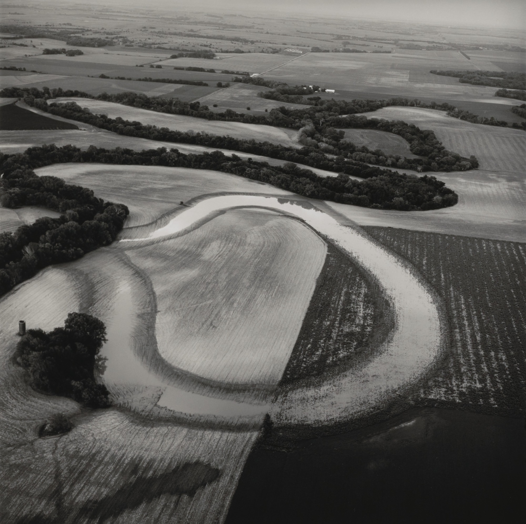 Aerial black-and-white photograph of a winding river creating an oxbow formation through agricultural farmland, with curved fields showing distinct plow patterns and tree-lined banks contrasting against lighter cultivated areas.