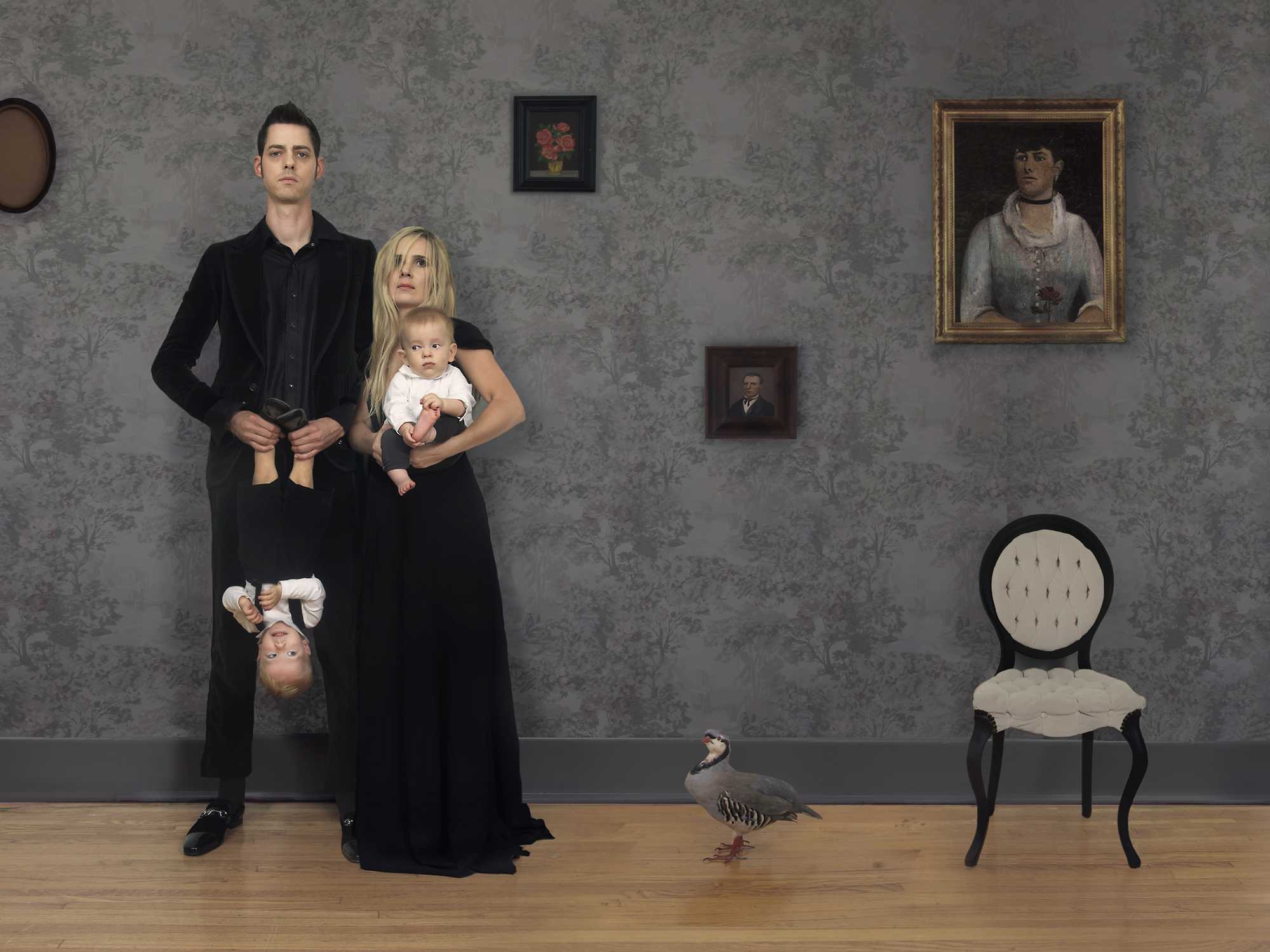 A man and woman in black clothing stand against a damask-patterned wall, with the woman holding two infants. A child hangs upside down from the man's arm, and a small goose walks across the wooden floor in the foreground. Period portraits and a round-backed chair are mounted on the wall behind them.