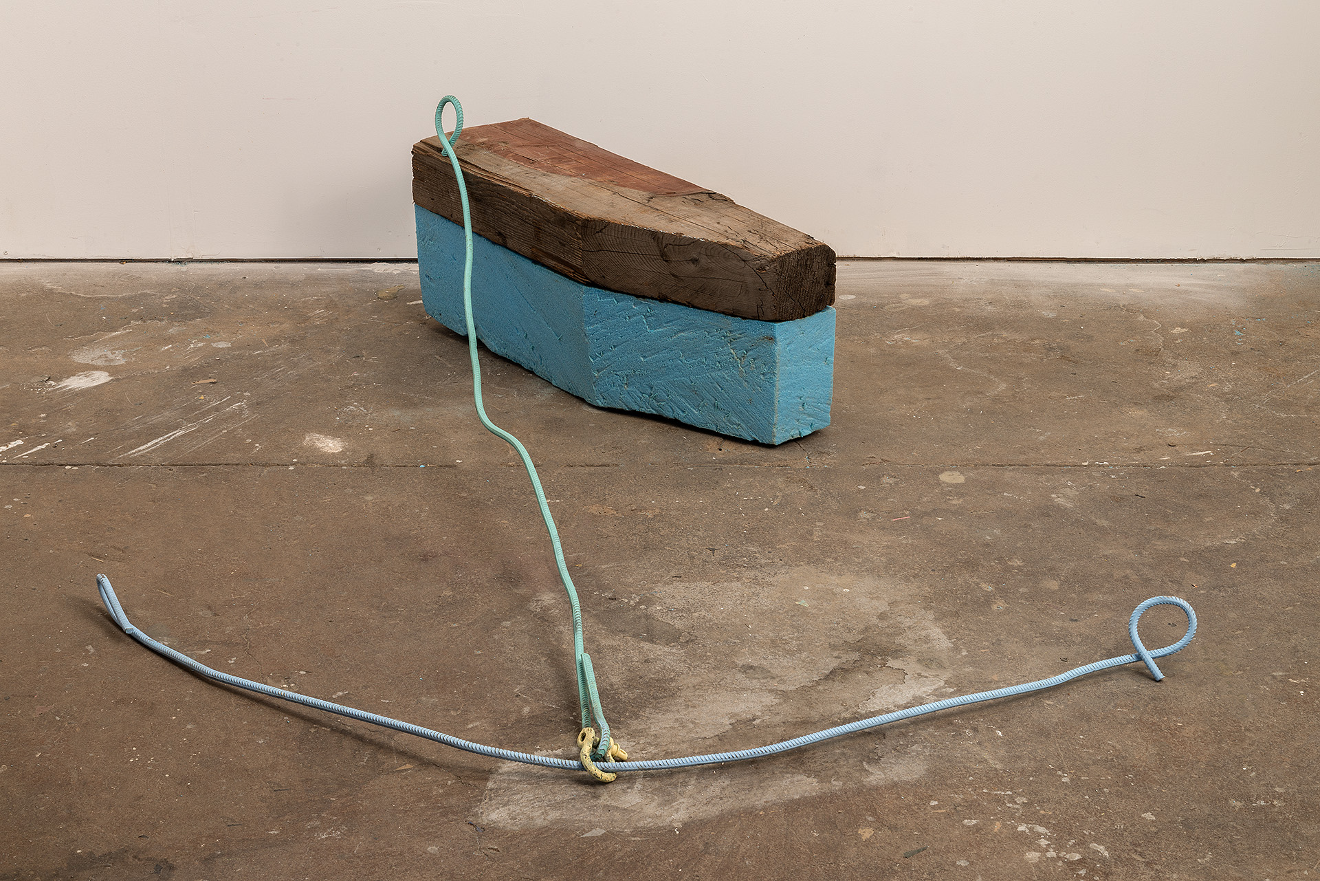 A turquoise-painted polystyrene block topped with weathered wood sits on a concrete floor, anchored by two lengths of blue metal cable with looped ends extending across the ground.