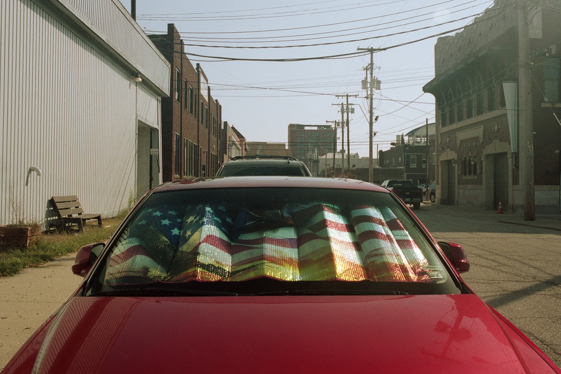A red car parked in an industrial alleyway displays an American flag sunshade across its windshield, with brick and corrugated metal buildings lining the narrow street under overcast skies.