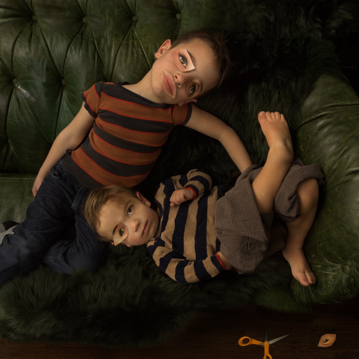 Two young boys lie on a green quilted upholstered surface, viewed from above. The older boy wears a red and black striped shirt, while the younger wears a tan and navy striped sweater. Both gaze upward toward the camera with contemplative expressions. A pair of orange scissors and a small diamond-shaped object rest on a dark wooden surface at the bottom of the frame.