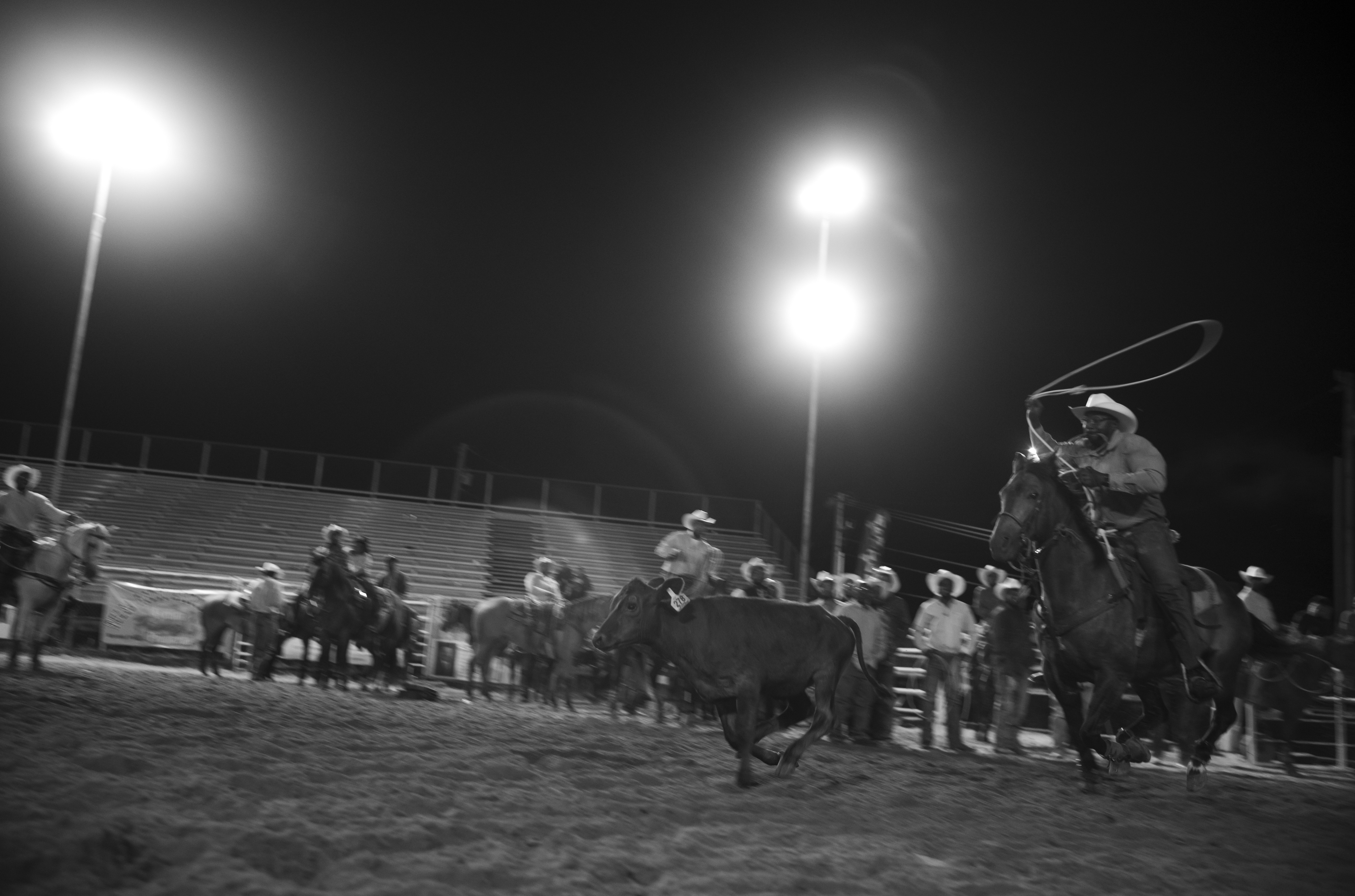 Black and white photograph of a nighttime rodeo scene lit by bright floodlights. A cowboy on horseback swings a lasso in the arena while spectators watch from the stands and ground level, their figures silhouetted against the dark sky.