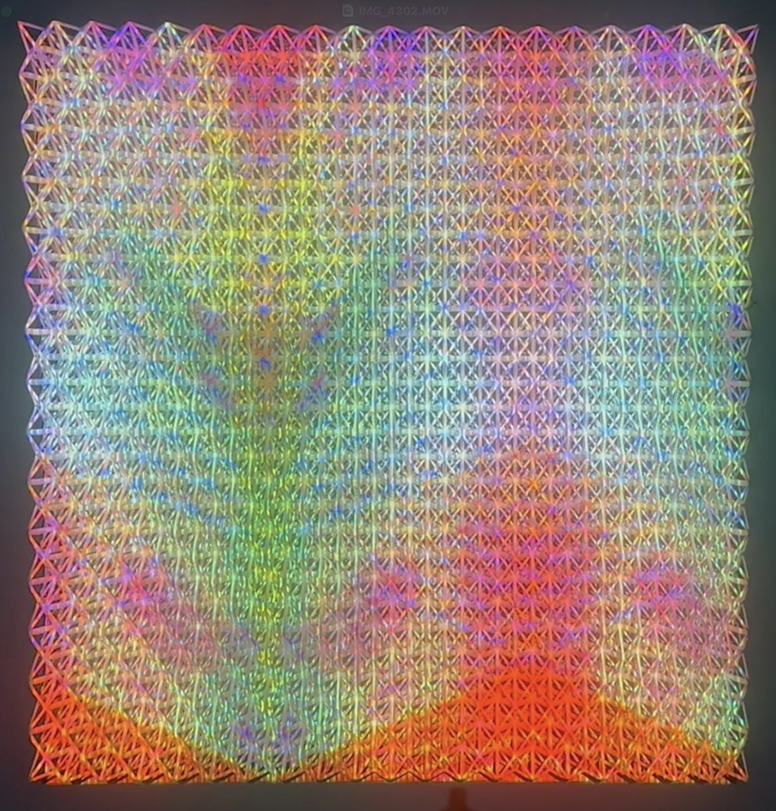 A large square composition of densely packed triangular forms creating an intricate lattice pattern. The work features a vivid color gradient transitioning from cool purples and blues in the upper left through vibrant greens in the center to warm oranges and reds in the lower right, with subtle iridescent and reflective qualities throughout.