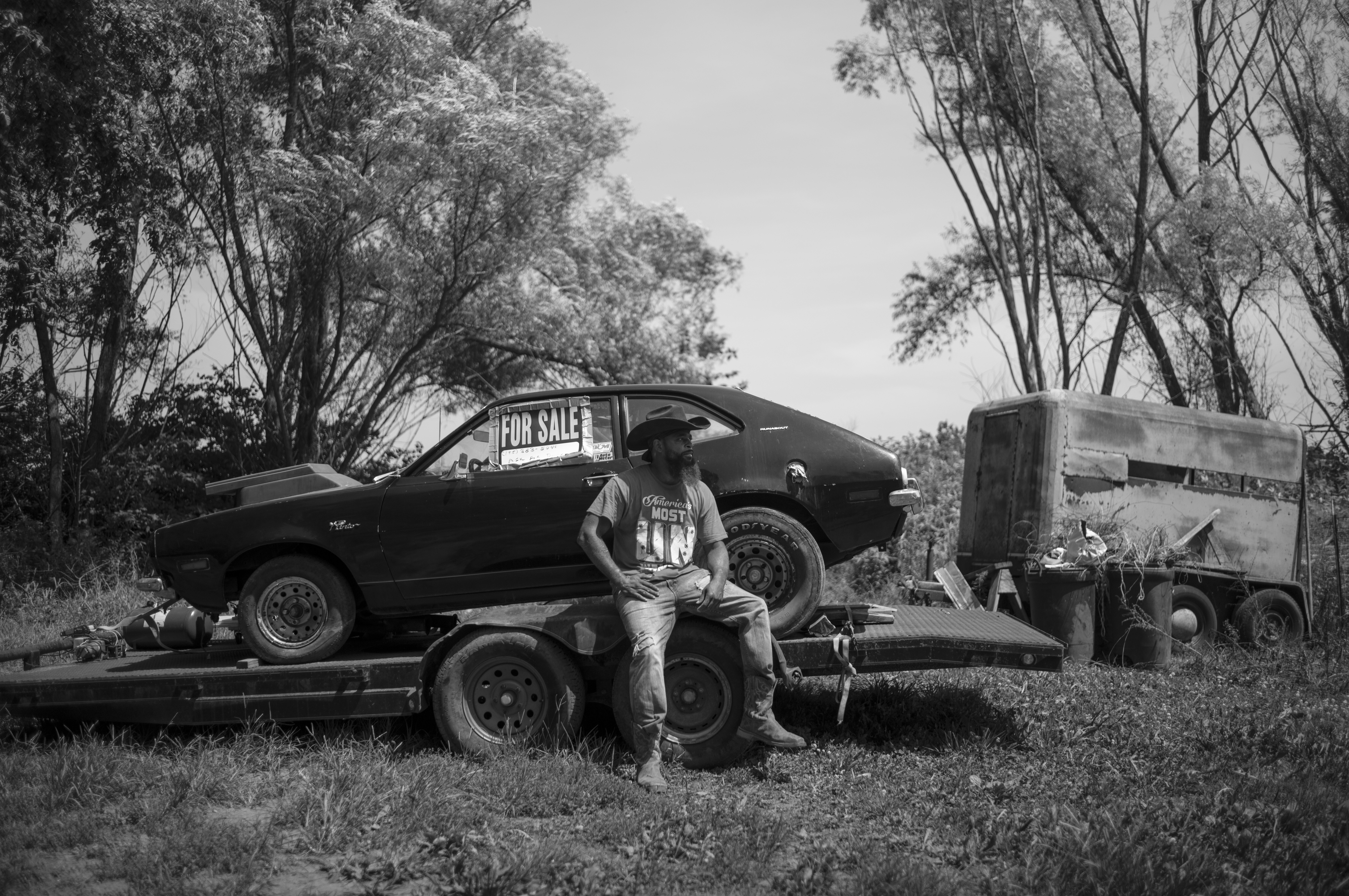 Black and white photograph of a man sitting on a flatbed trailer in a rural setting, wearing a baseball cap and t-shirt. Behind him sits a vintage car with a "For Sale" sign displayed in its window, positioned on the trailer. Overgrown vegetation and bare trees frame the composition, with an abandoned truck visible in the background, creating a desolate landscape narrative.