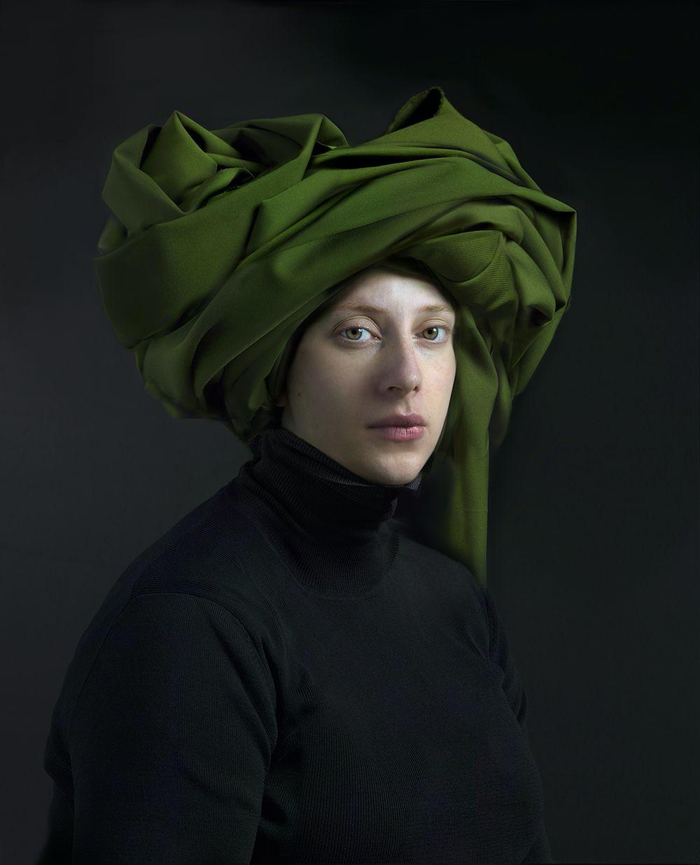 Portrait of a pale-skinned person wearing a black turtleneck and a voluminous olive-green fabric draped and wrapped around their head like a turban, photographed against a dark gray background with directional lighting that emphasizes the sculptural folds of the fabric.