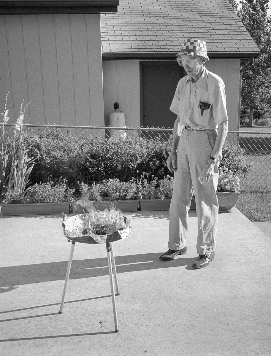 Black and white photograph of an older man in light-colored work clothes and a checkered hat standing beside a tripod-mounted grill or fire bowl with burning coals and vegetation, with modernist residential architecture and planted gardens visible in the background.