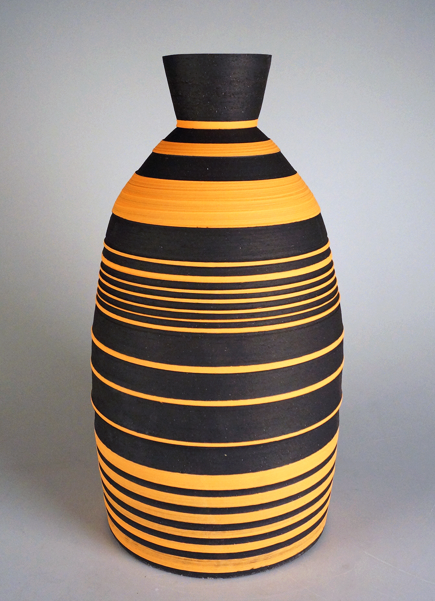 A tall, tapered ceramic vase with a narrow cylindrical neck widens toward a rounded bulbous body, decorated with alternating horizontal stripes of black and bright orange-yellow that increase in frequency toward the base.