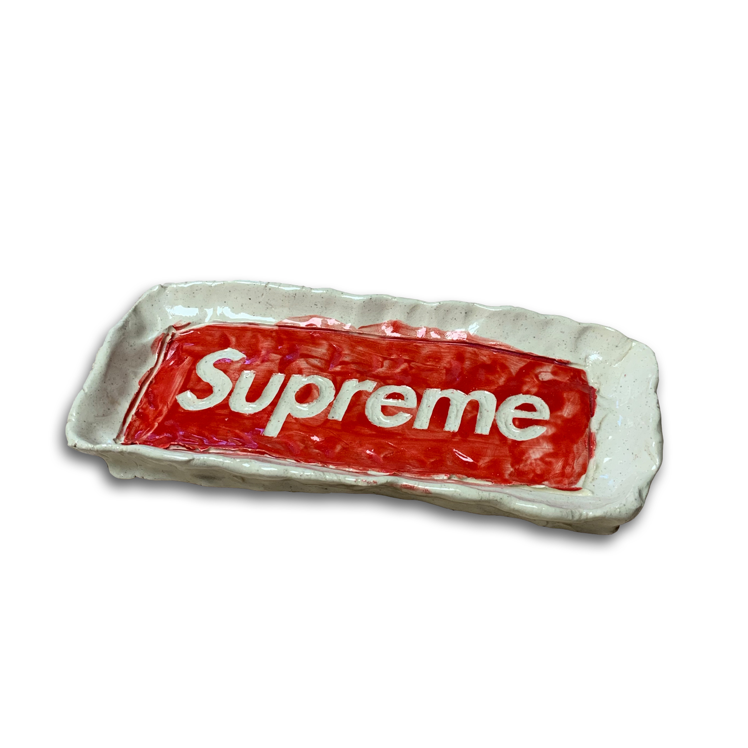 A ceramic ashtray with a speckled cream-colored glaze features a bold red rectangular form glazed with white lettering reading "supreme" across its center. The piece sits within the shallow clay vessel with organic, slightly irregular edges.