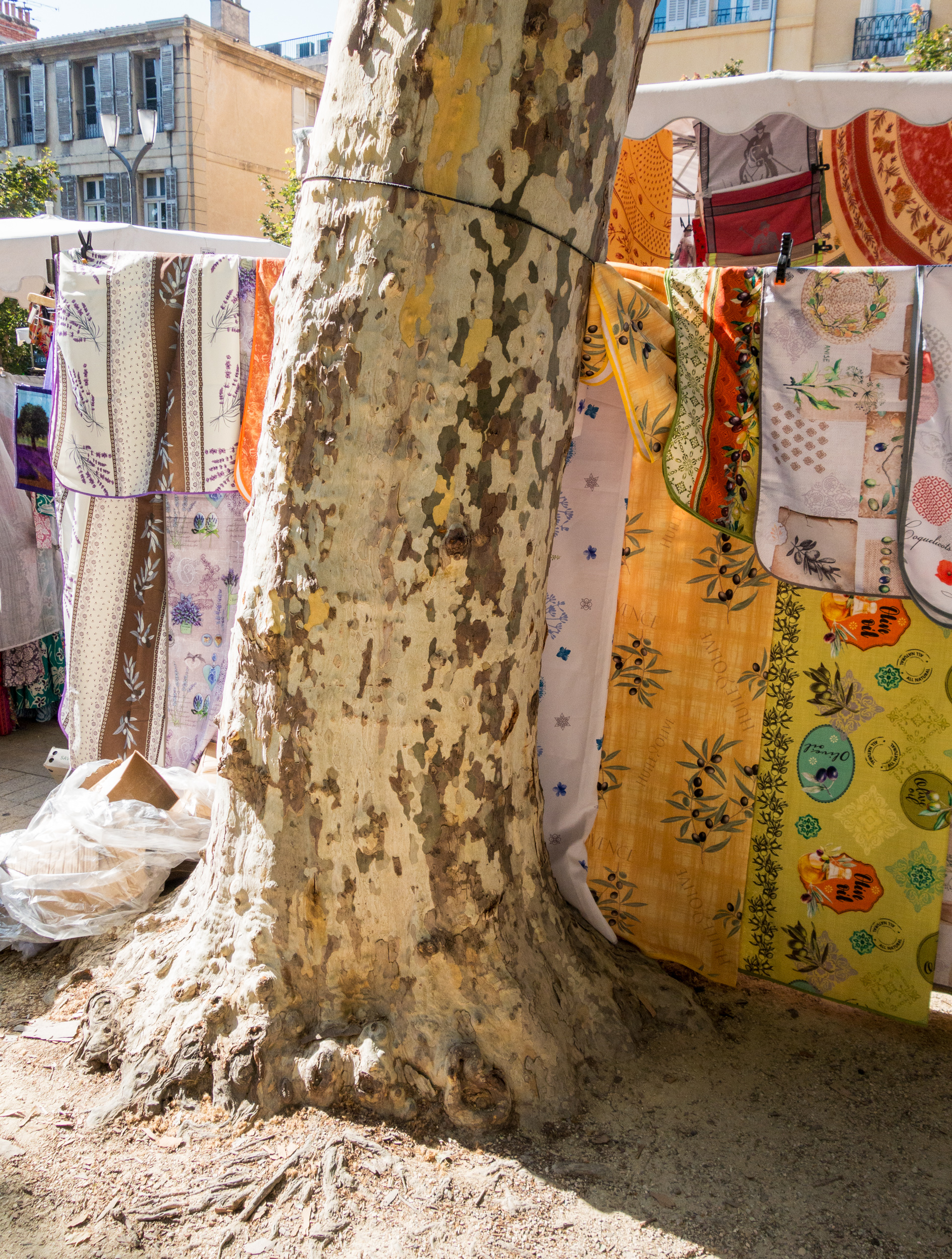 Colorful fabrics and textiles hang on clotheslines strung between a weathered, cream-colored tree trunk and white structures in a sunlit Mediterranean courtyard. Patterned cloths in shades of orange, yellow, cream, and burgundy with floral and geometric designs dry in the shade, while pastel-colored buildings are visible in the soft-focused background.