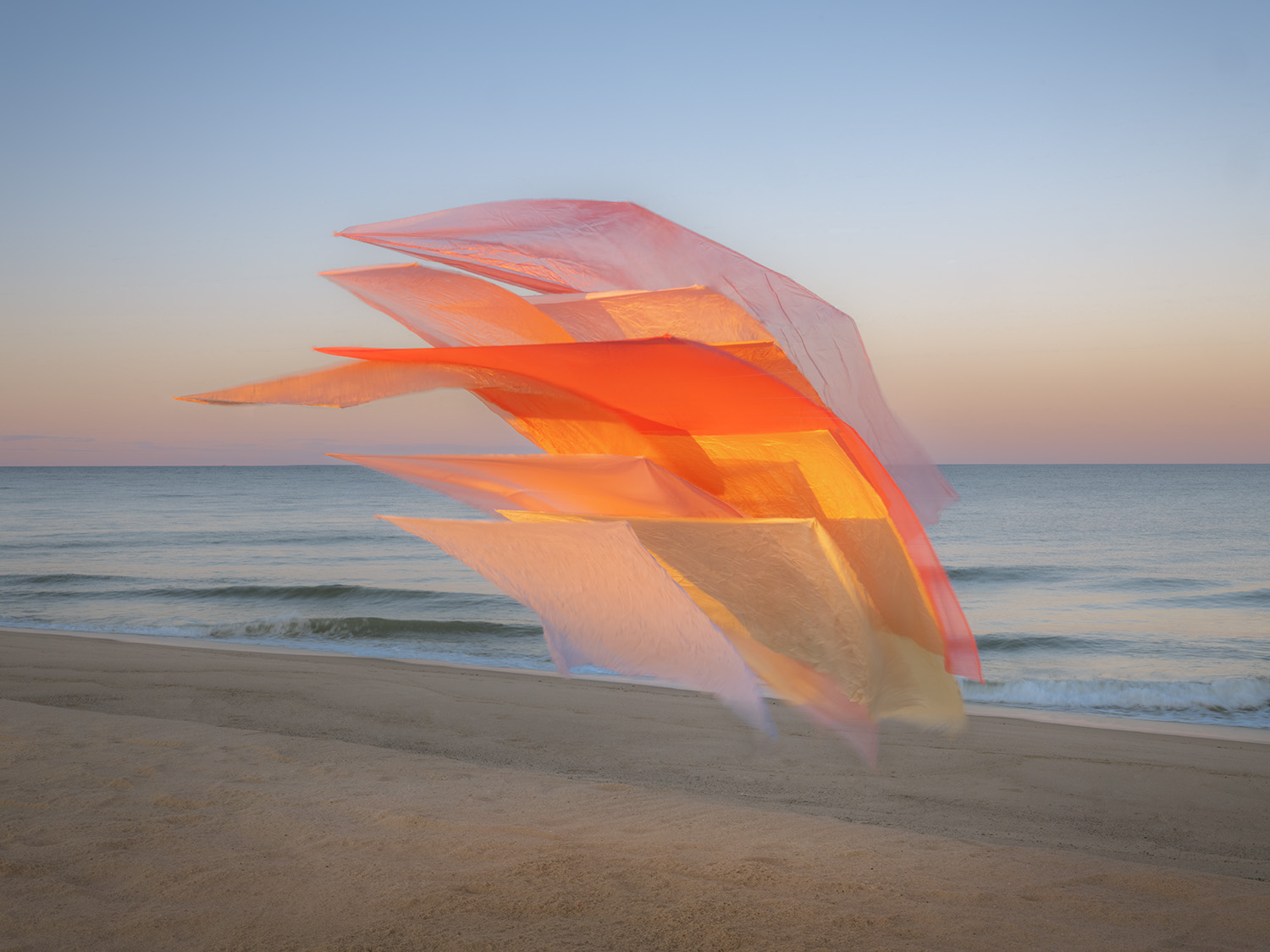 Layered sheets of translucent silk in graduated shades of pink, coral, orange, and yellow billow in the wind on a beach at dusk, creating an ethereal sculptural form against the calm ocean and twilight sky.