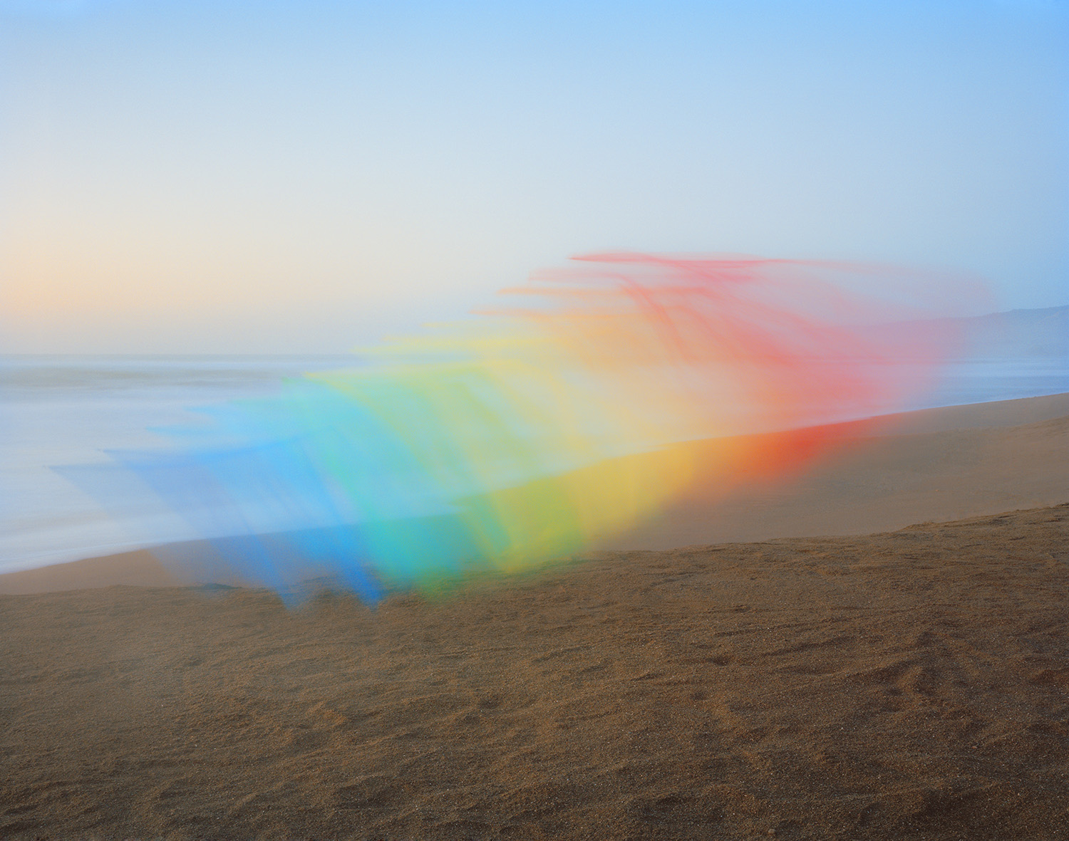 A rainbow gradient of tulle fabric stretches diagonally across a misty beach landscape, its translucent layers transitioning from blue through green, yellow, orange, and pink against a pale dawn sky and sandy shore.