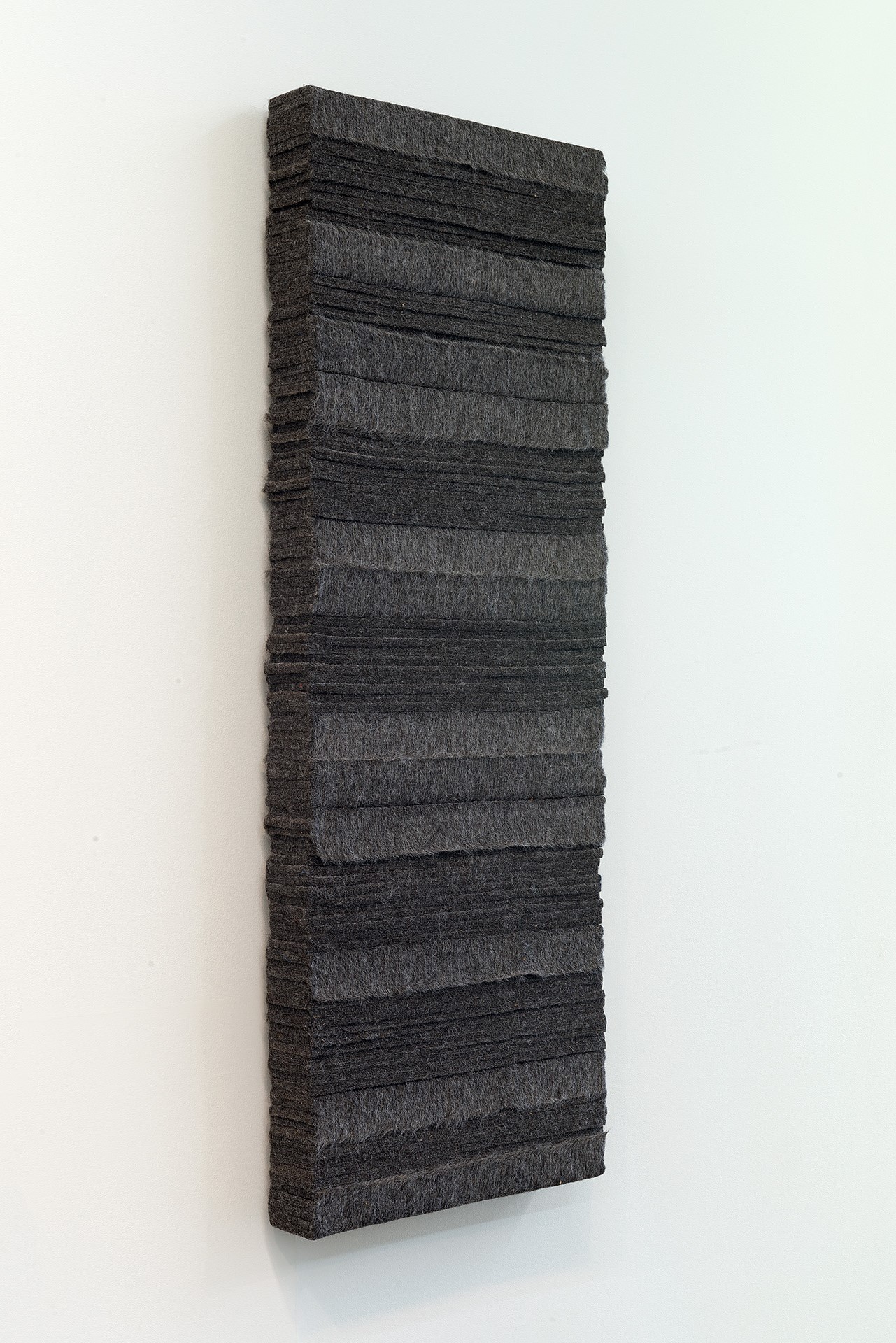 A tall rectangular panel of dark charcoal-colored industrial felt mounted vertically on a white wall, with horizontal bands of varying widths creating subtle texture and tonal shifts across its surface.