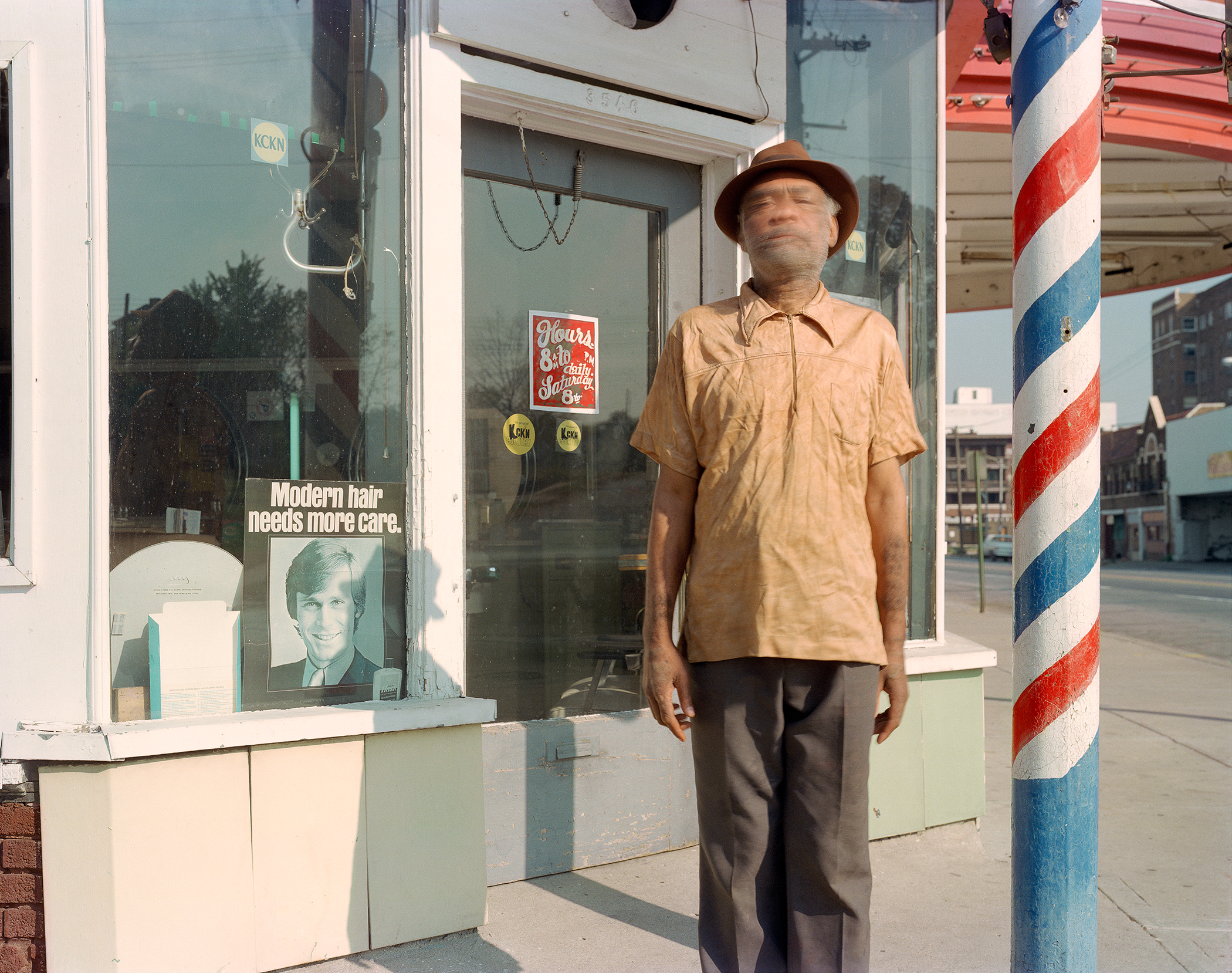 A man wearing a tan short-sleeved shirt and brown pants stands on a city sidewalk in front of a barber shop, wearing a brown hat and looking directly at the camera. A distinctive red, white, and blue barber pole is visible to his right, and the shop window behind him displays vintage hair care advertisements and signage.