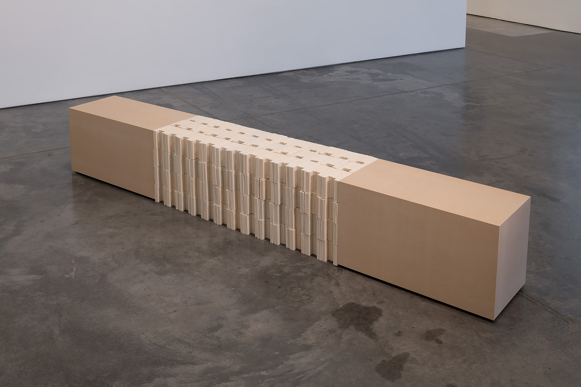 A long, low horizontal sculpture made of cream-colored material spans across a dark concrete floor. The center section features a dense accordion-like arrangement of thin vertical elements, creating a rhythmic linear pattern, while solid rectangular blocks anchor each end.