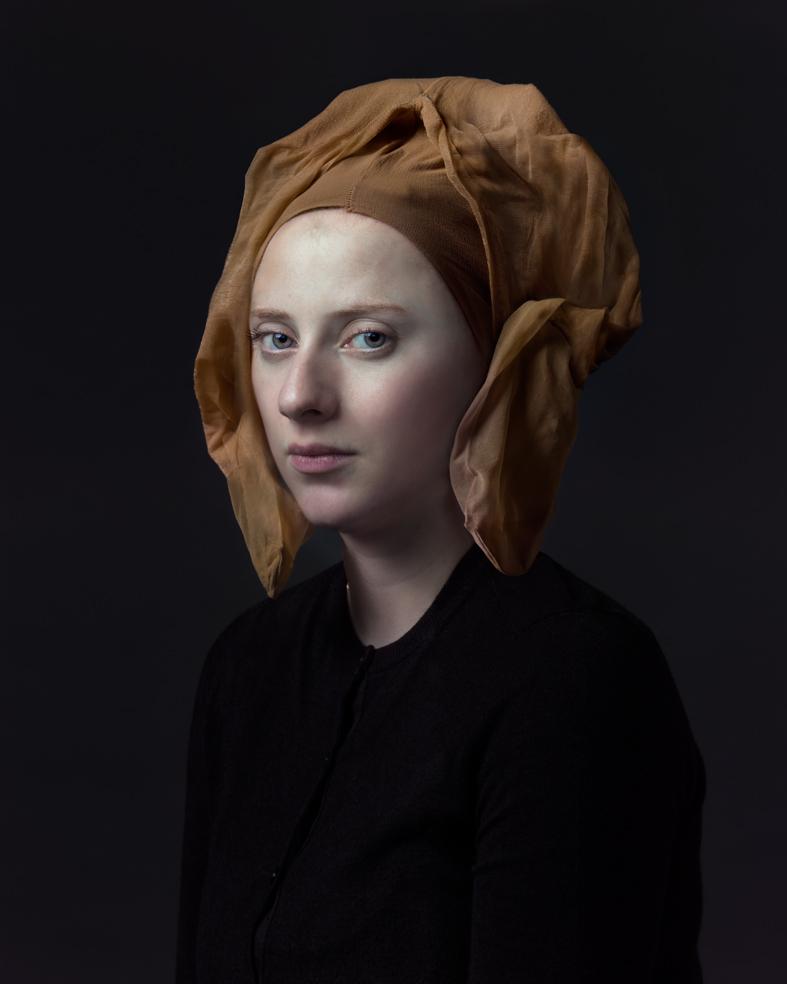 Portrait of a pale-skinned figure with a shaved head wearing a tan linen headwrap and black garment, photographed against a dark background with subtle lighting that emphasizes the sculptural quality of the face and the textural folds of the fabric.