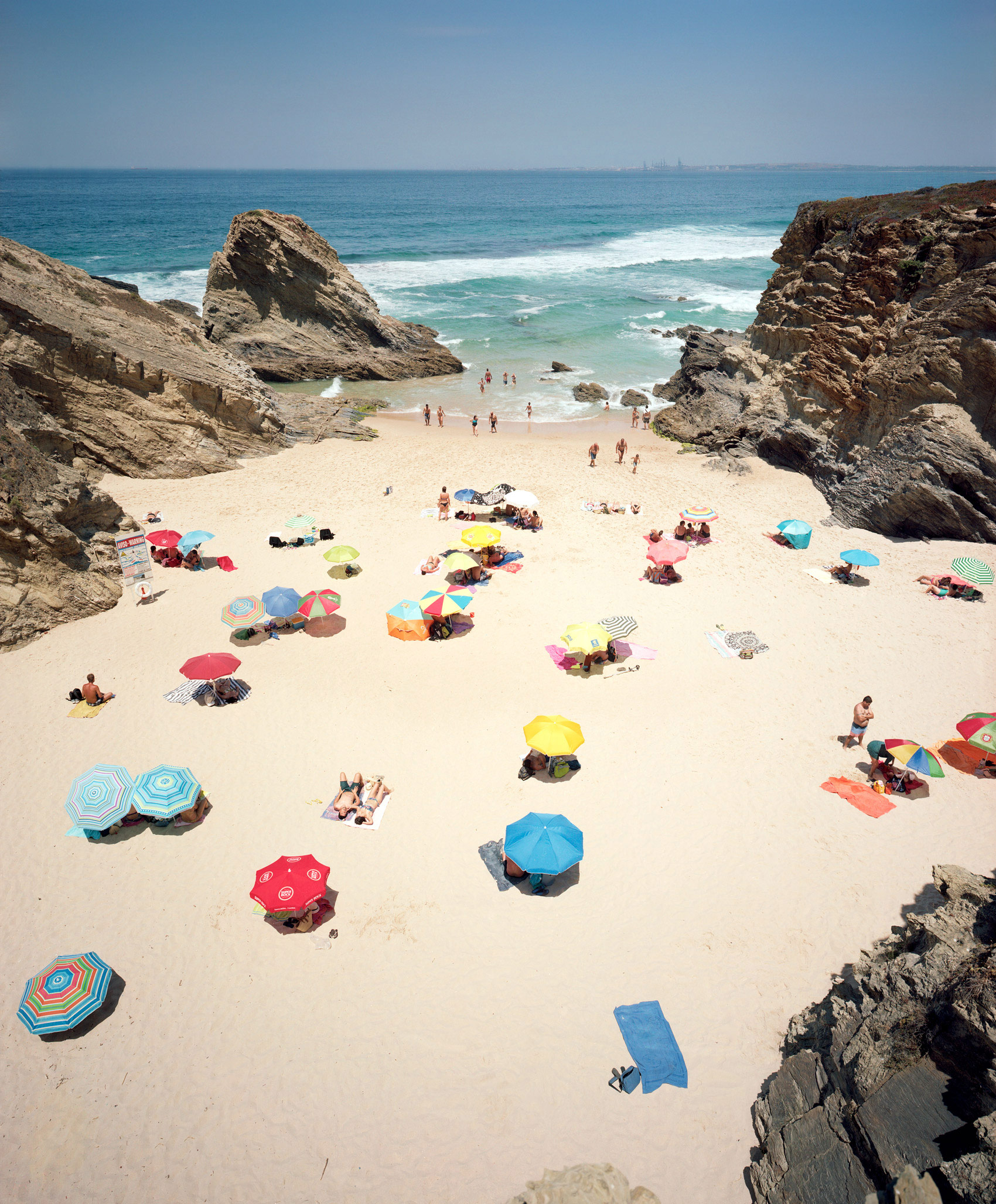 Aerial view of a sandy beach cove framed by dramatic rocky outcrops, dotted with colorful umbrellas in red, blue, yellow, and striped patterns. Beachgoers scatter across the pale sand while waves break along the shoreline, with a calm blue ocean and distant horizon visible beyond.