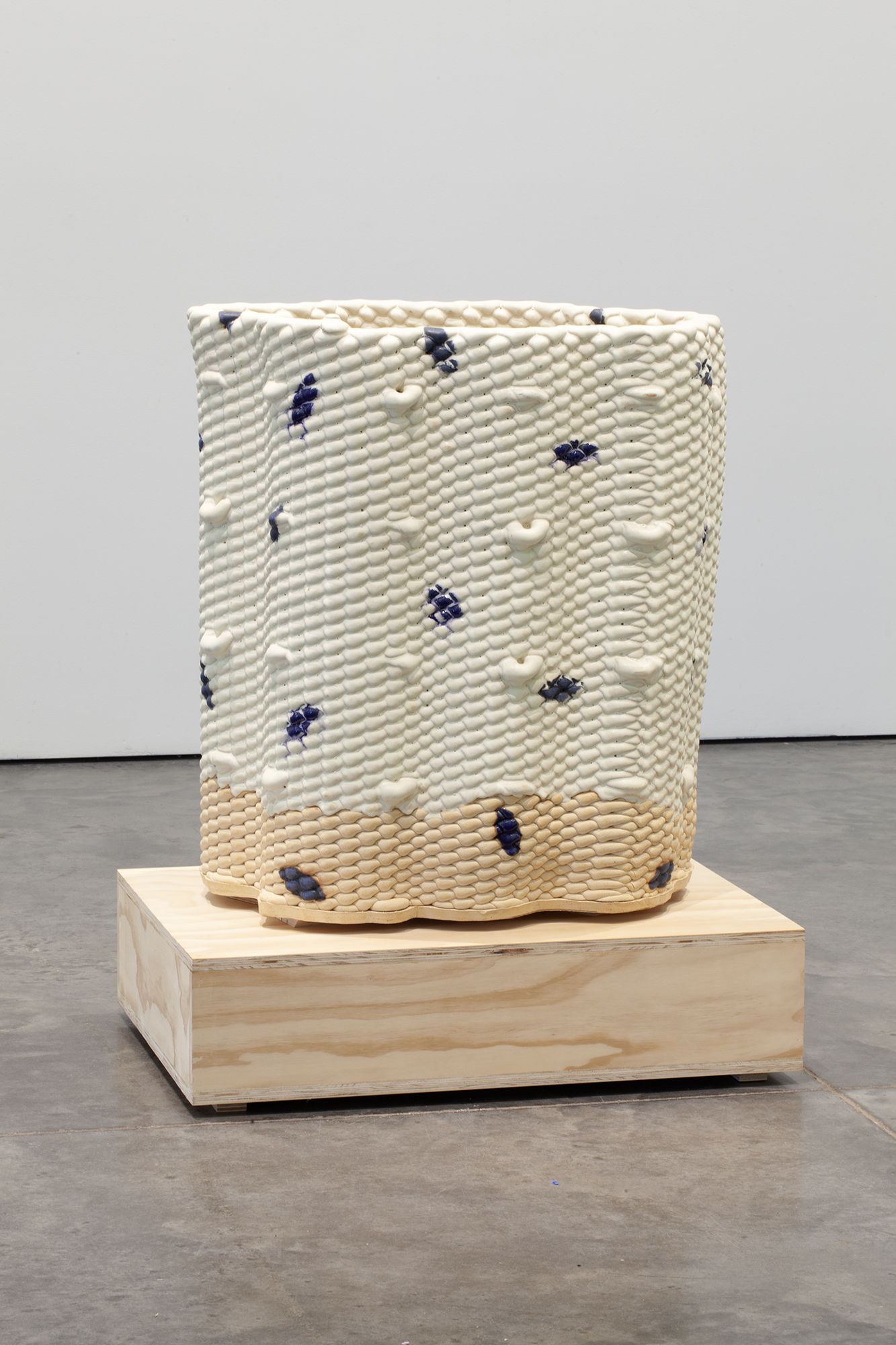 A cylindrical ceramic vessel with a textured, bumped surface in cream and pale yellow tones, scattered with dark blue glazed marks. The piece sits on a light wooden base against a gallery floor and white wall.