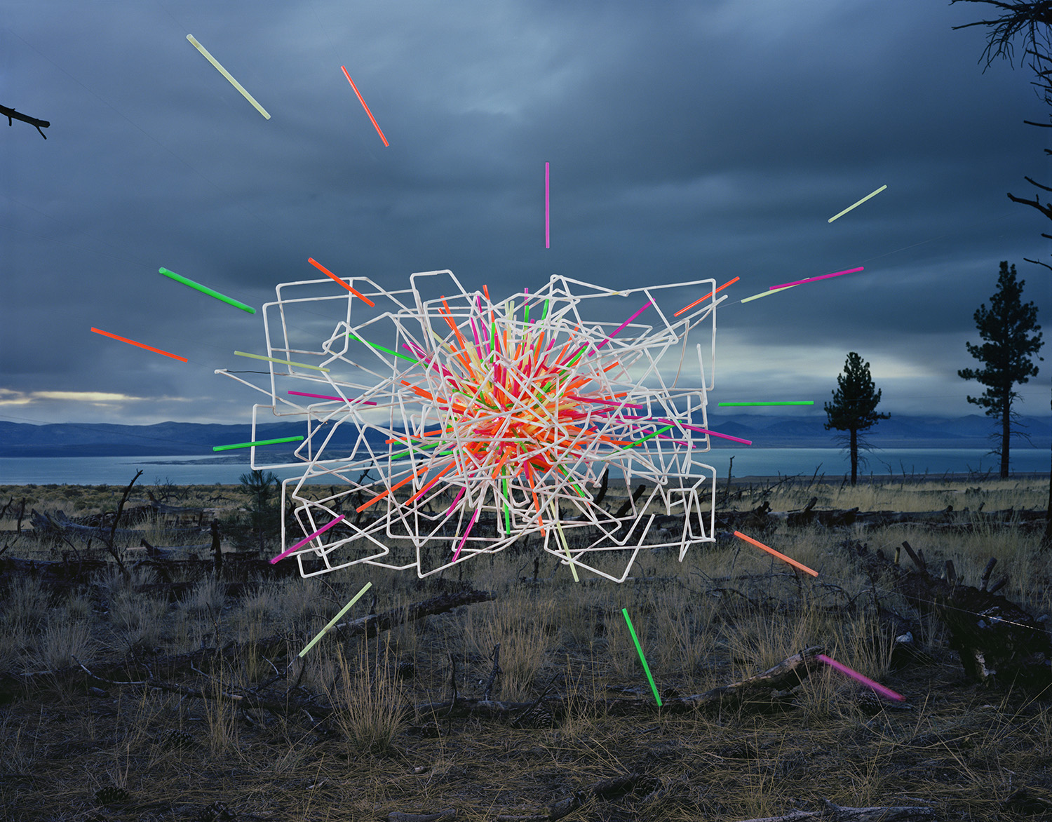 Neon-colored straws and white geometric wire frames arranged in a chaotic sculptural composition float above a desert landscape with a large lake visible in the distance under a dramatic cloudy sky.