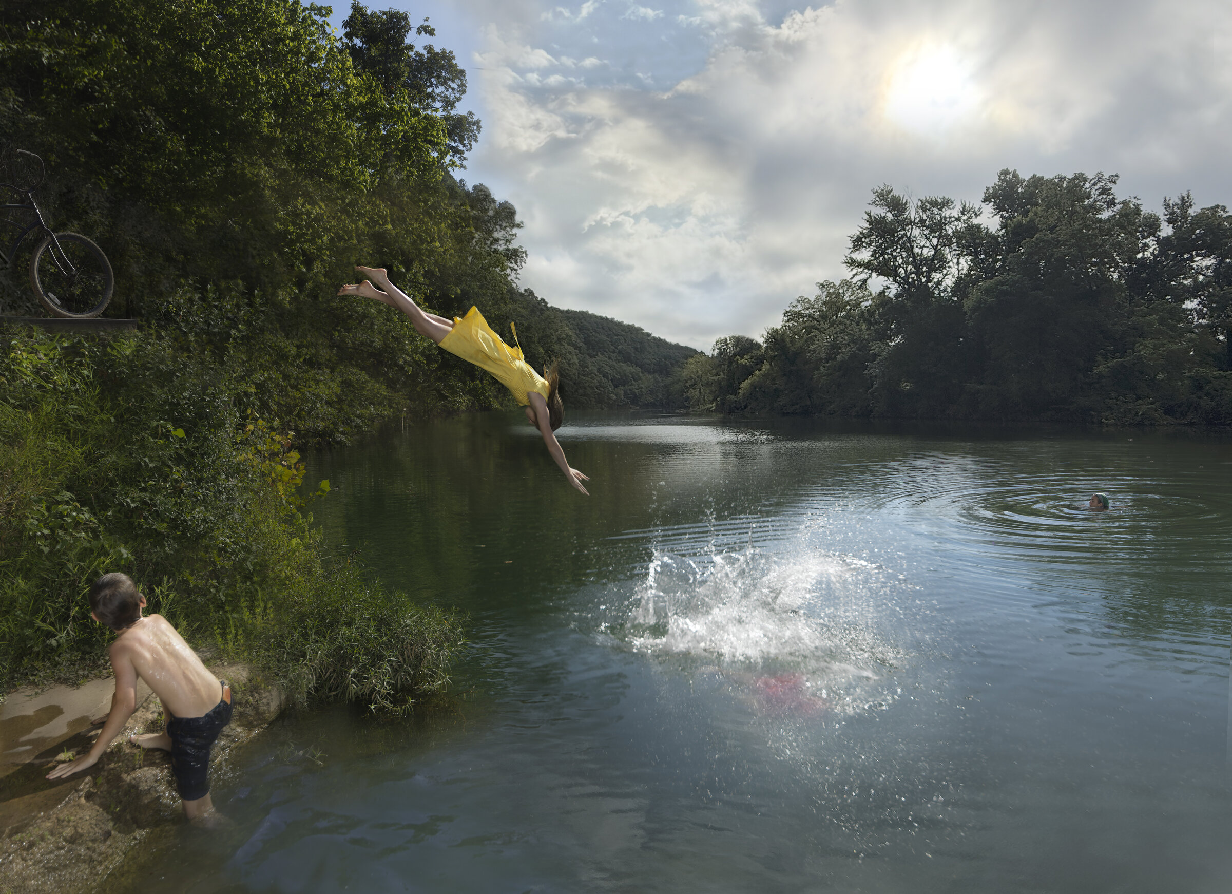A person in a bright yellow shirt dives horizontally from vegetation along a riverbank toward calm water, while another figure wades in the shallows nearby. Forested hills and a cloudy sky frame the serene landscape, with a bicycle visible in the upper left corner.