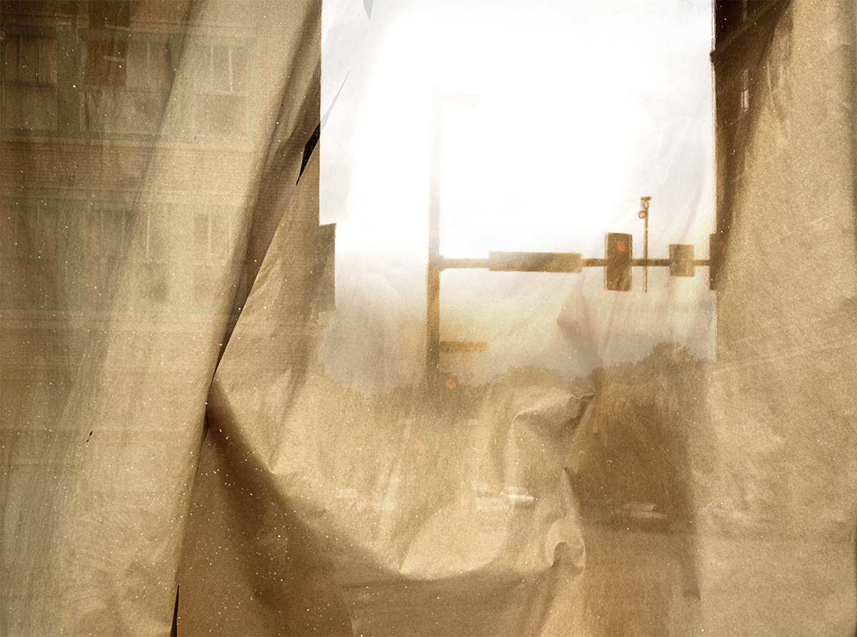 Translucent layered paper creates a hazy, atmospheric composition in warm beige and cream tones. A rectangular opening frames an indistinct industrial structure silhouetted against bright light, with dust or particles suspended throughout the composition.