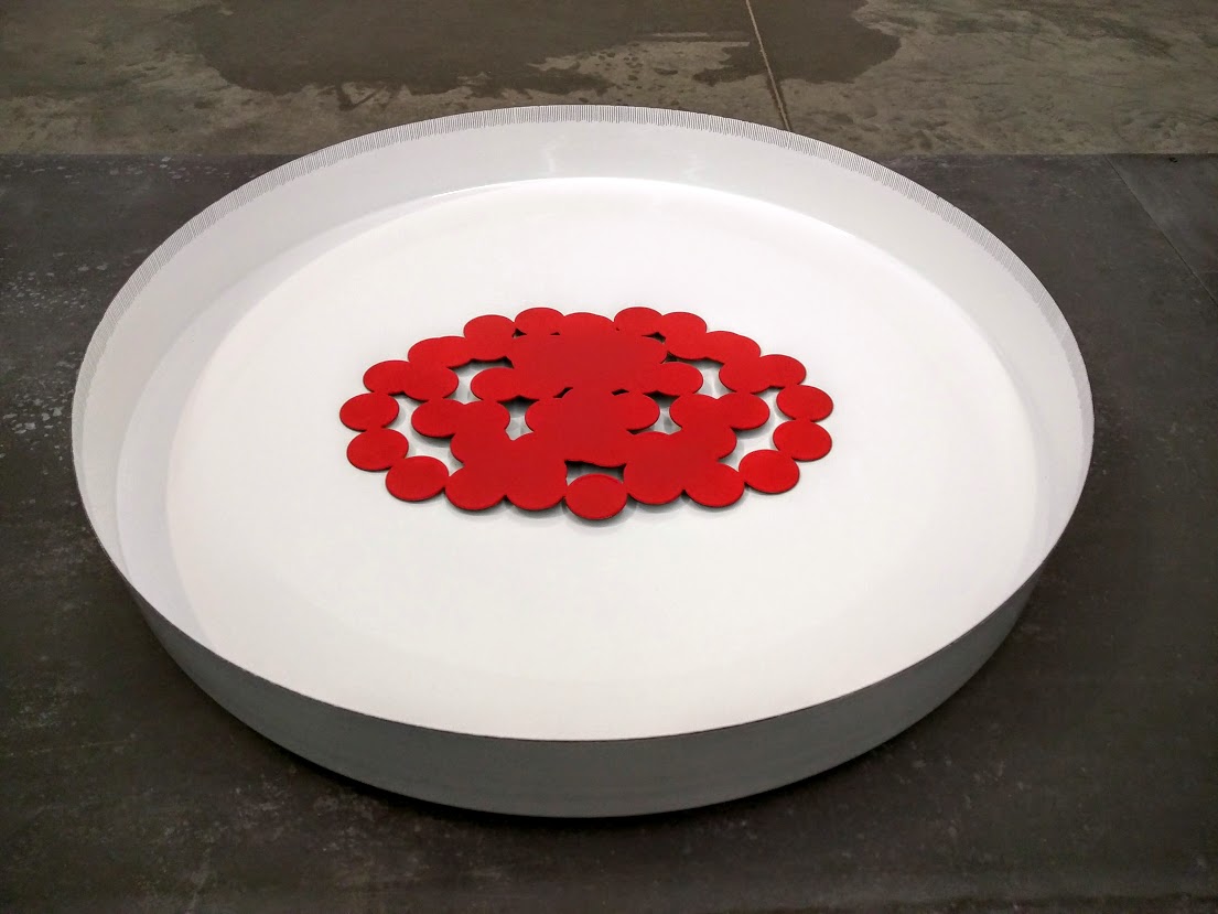A large white circular shallow bowl contains a cluster of red circular forms arranged in the center, with algorithm-generated line drawings etched into their surfaces, creating an intricate pattern against the stark white background.