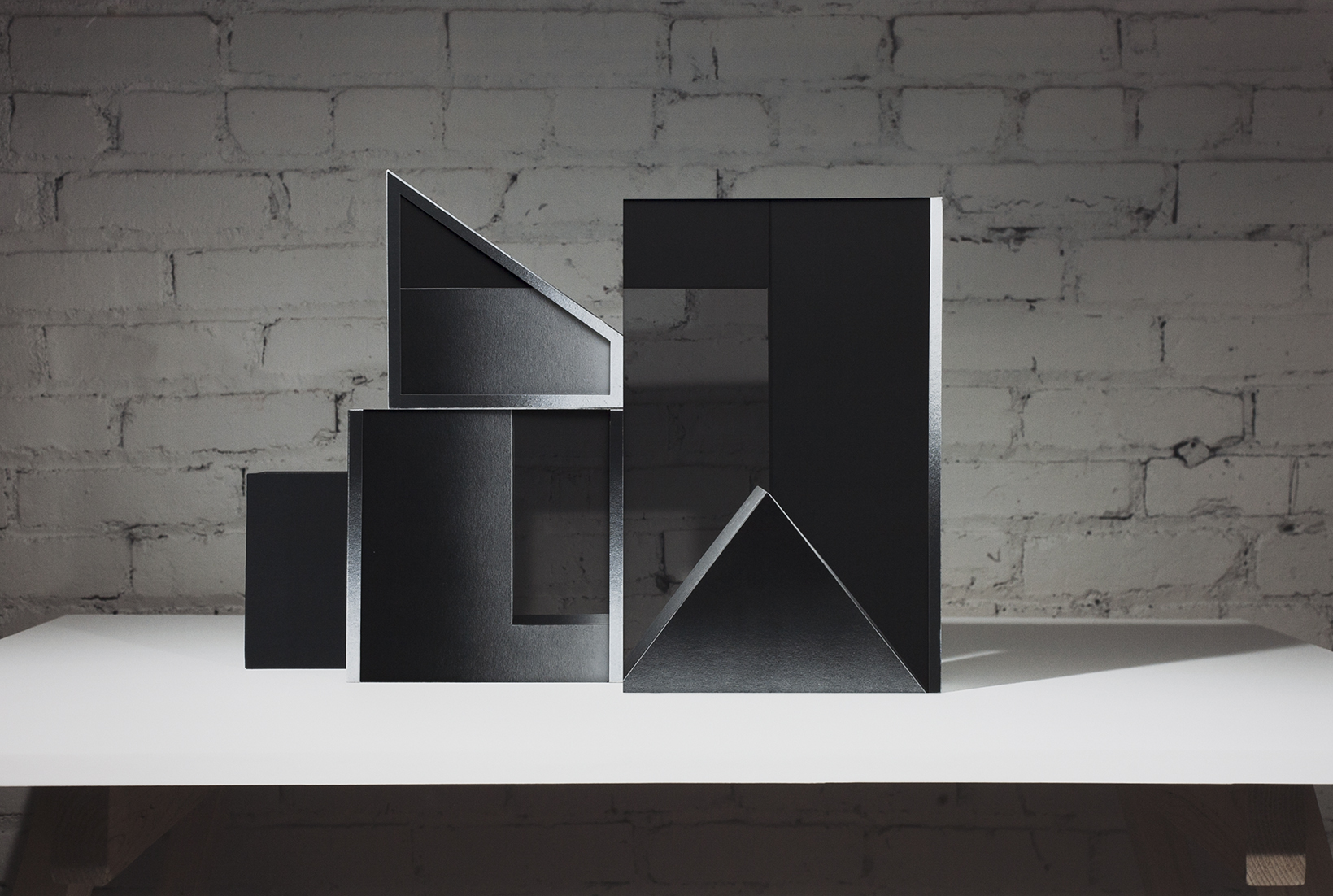 Abstract geometric sculpture made of black layered materials arranged in ascending rectangular and triangular forms on a white platform. The composition features sharp angles, cutouts, and overlapping planes that create dramatic shadows against a weathered brick wall.