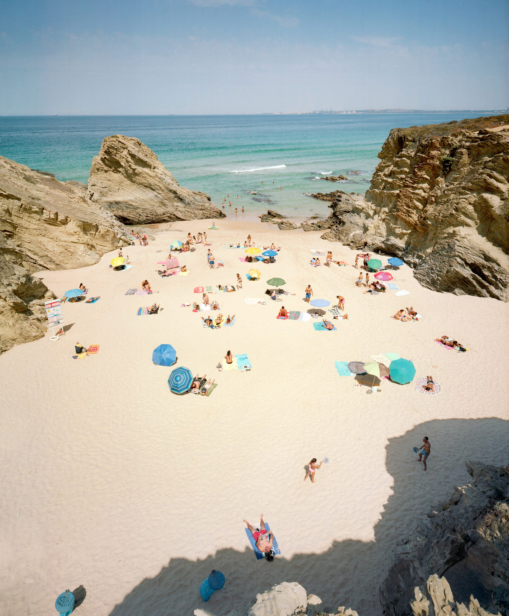 Aerial view of a crowded sandy beach nestled between two large rocky outcroppings, with beachgoers scattered across the sand beneath colorful umbrellas and towels, turquoise water and gentle waves visible beyond.