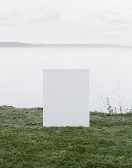 A large white rectangular form stands centered in a grassy field overlooking a calm body of water and distant shoreline under an overcast sky. Sparse dried vegetation and a leafless branch appear to the right of the geometric structure.