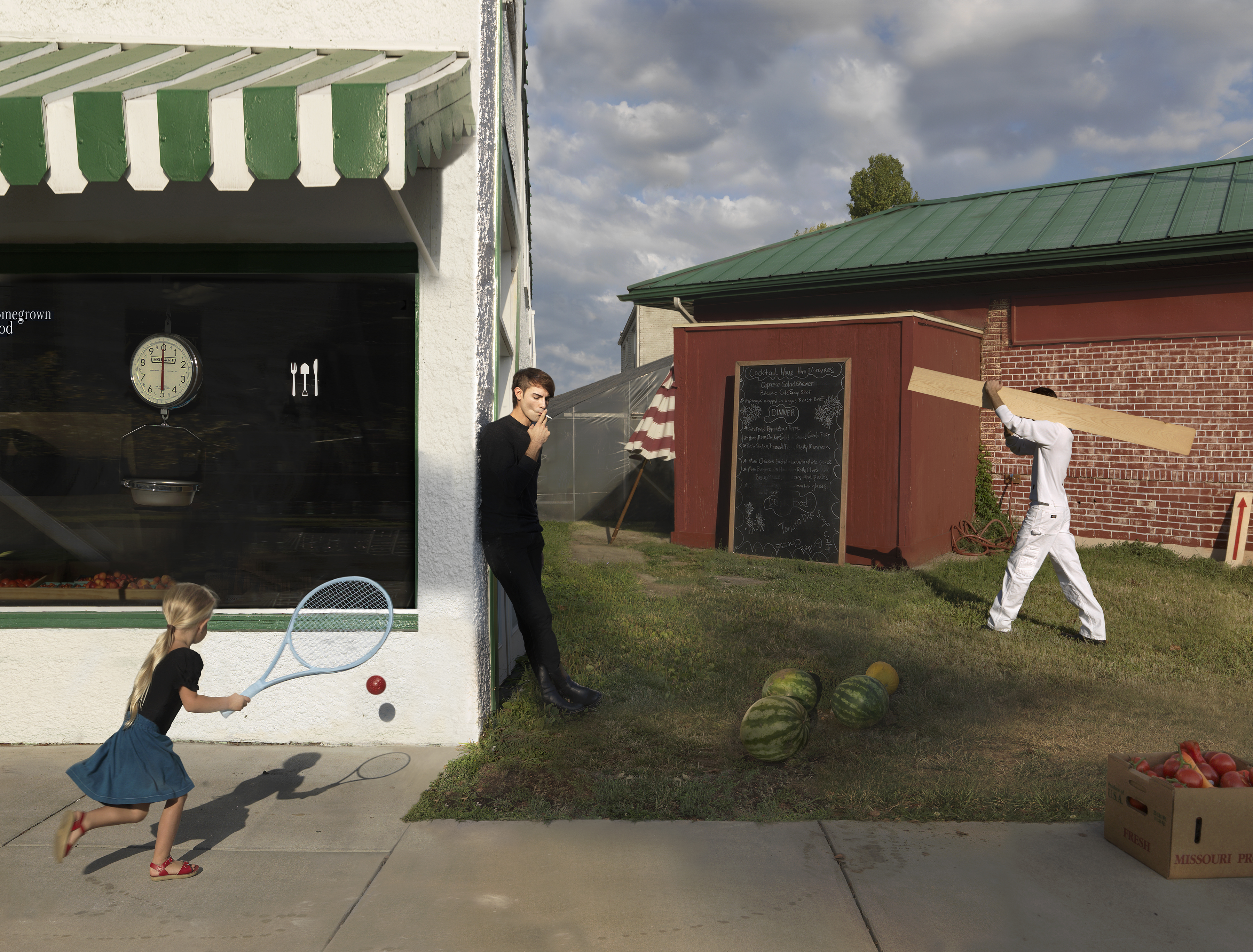 A surreal domestic scene depicting a farm building with green and white striped awning where a young girl in a blue skirt plays tennis against the storefront window. A man in dark clothing stands nearby observing, while a figure in white clothing swings an oversized baseball bat in a grassy yard. Melons and produce are scattered across the ground, with a box labeled "Missouri" visible in the foreground. A red brick barn with green metal roof anchors the background under a partly cloudy sky.